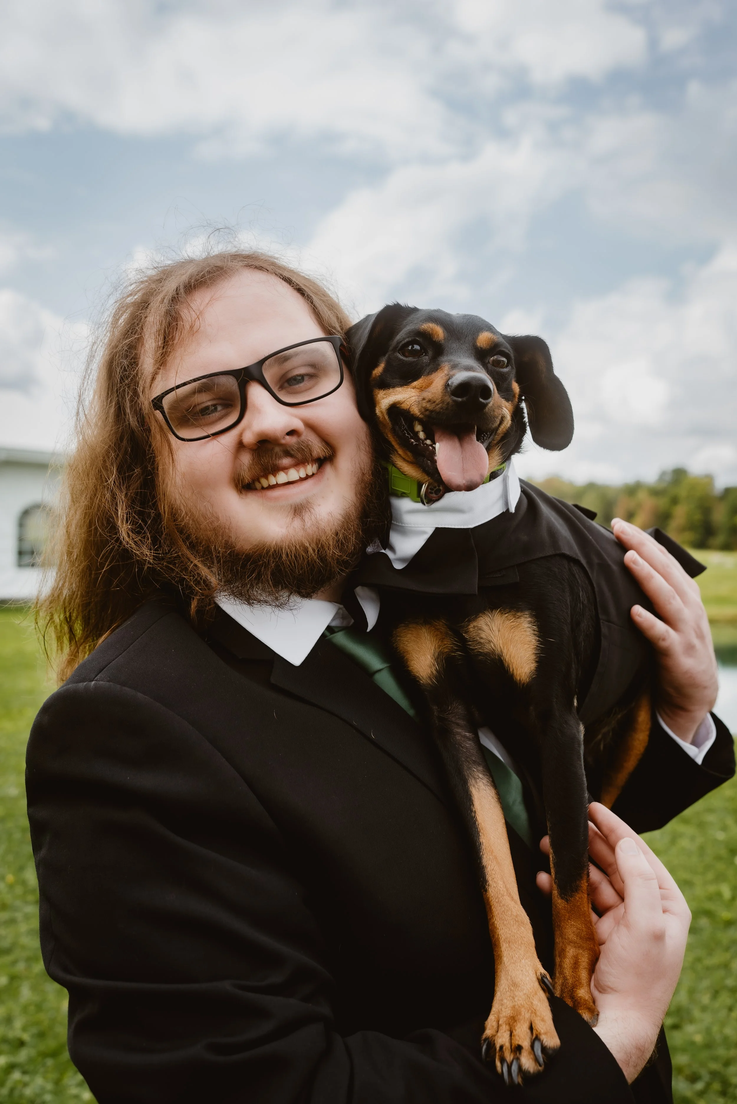 A man with glasses and long hair holding a happy black and tan dog outdoors on a cloudy day.