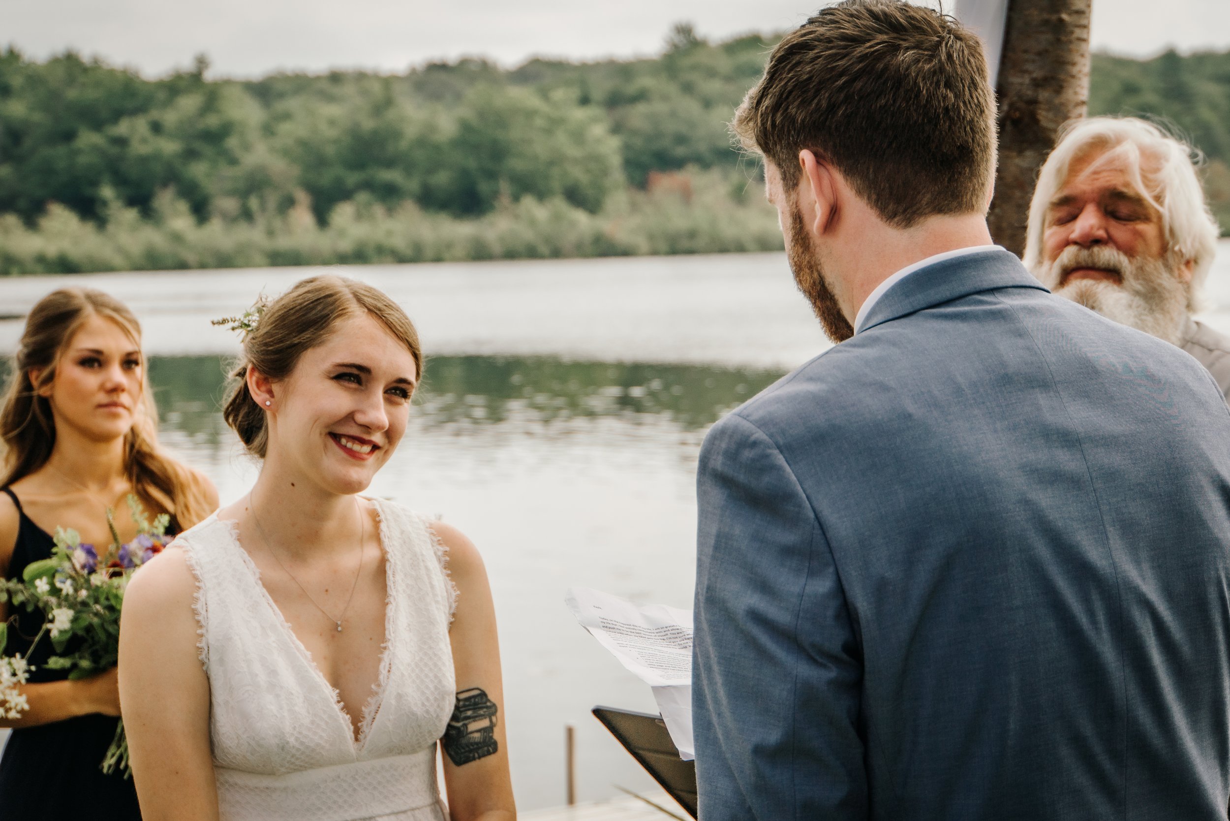 A wedding ceremony by a river with a couple exchanging vows, surrounded by friends and family, during daytime. Cassadaga, NY