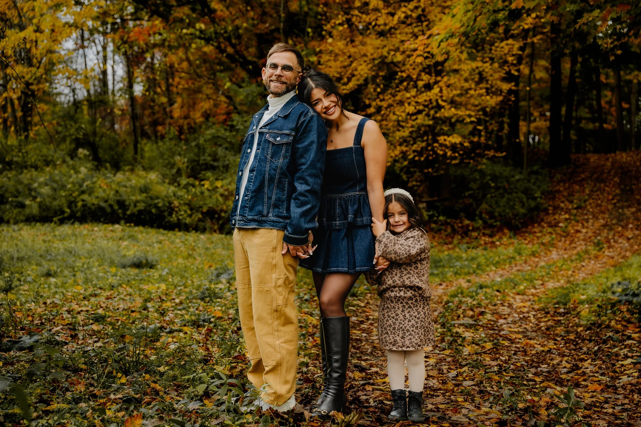 Family of three standing together outdoors in autumn, holding hands and smiling, with trees and fallen leaves in the background.