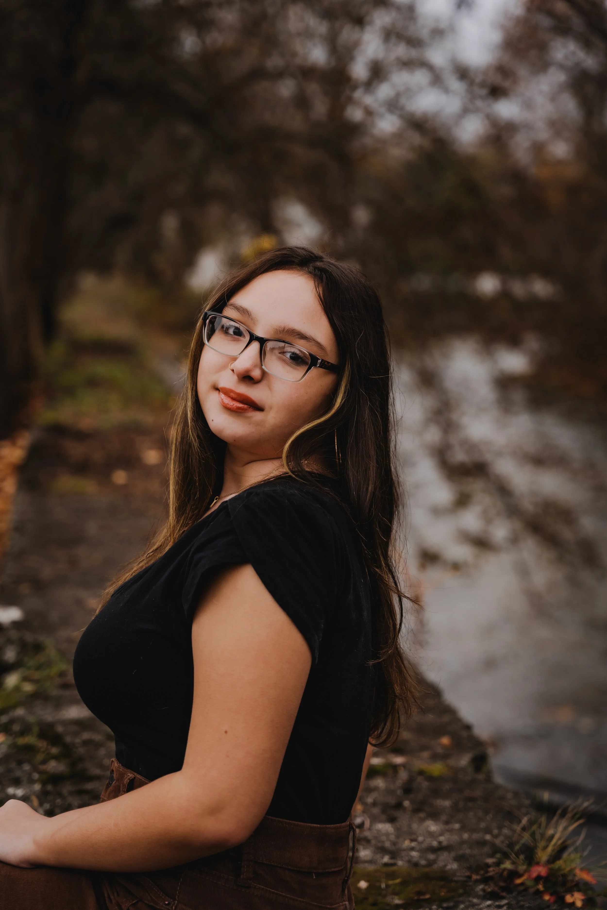 A woman with long dark hair and glasses standing near a river with trees in the background during fall. Fredonia, NY