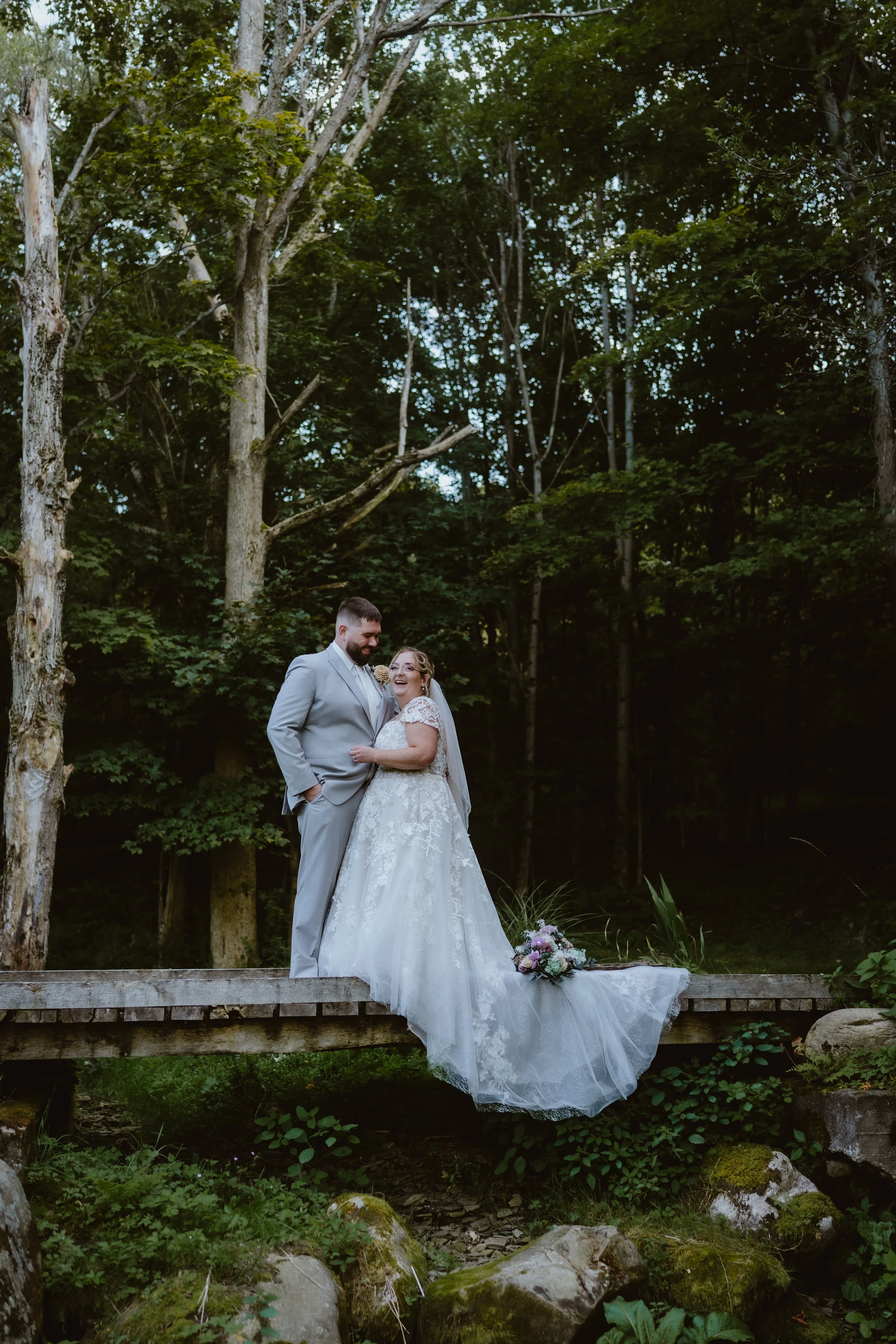 A bride and groom standing on a wooden bridge in a forest, smiling at each other. The bride is in a white wedding gown with lace details, and the groom is in a light gray suit. Farrington Hollow, Cherry Creek NY