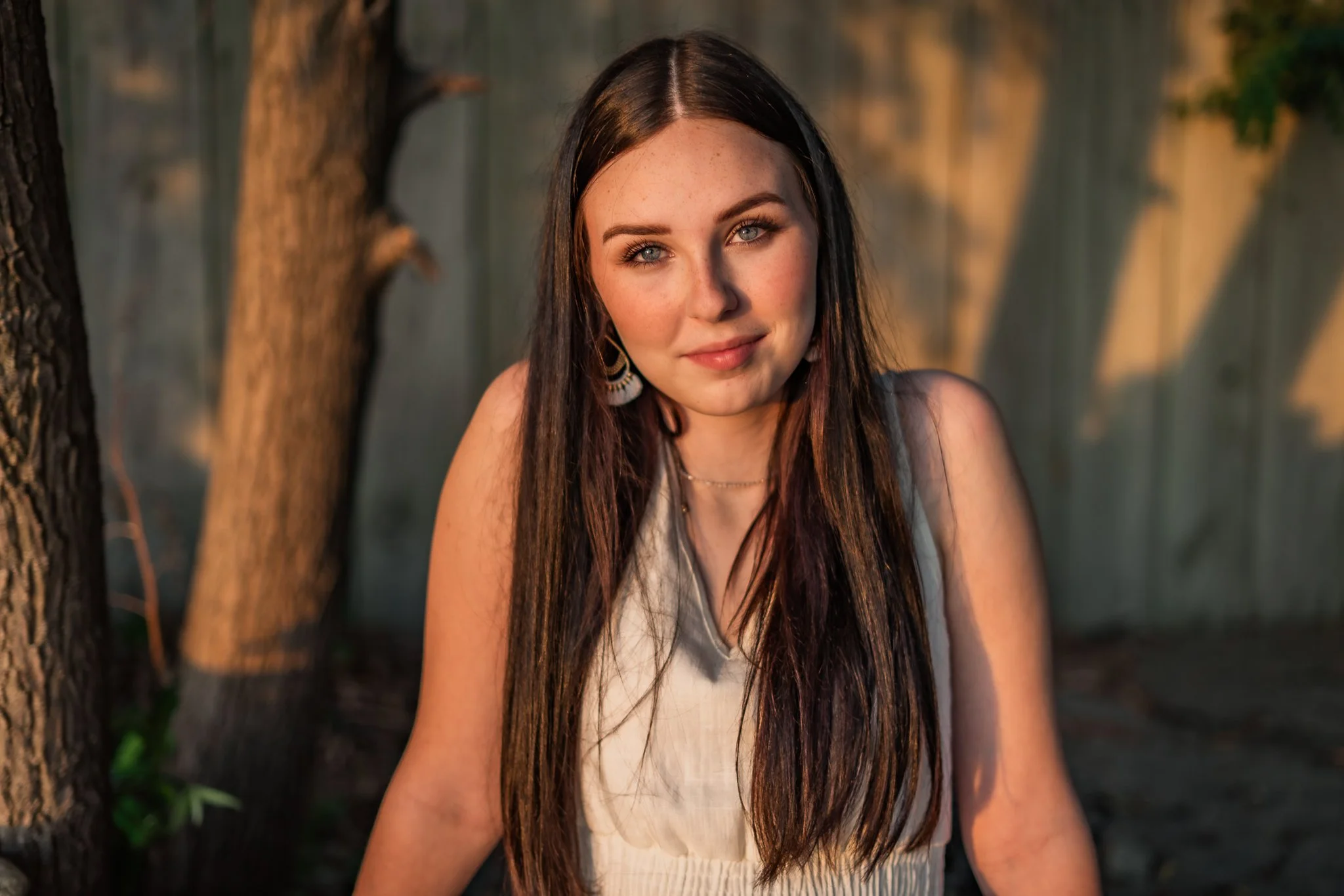Young woman with long dark hair and blue eyes outdoors near a tree and wooden fence, wearing a sleeveless beige top and jewelry, illuminated by warm sunlight. Westfield, NY