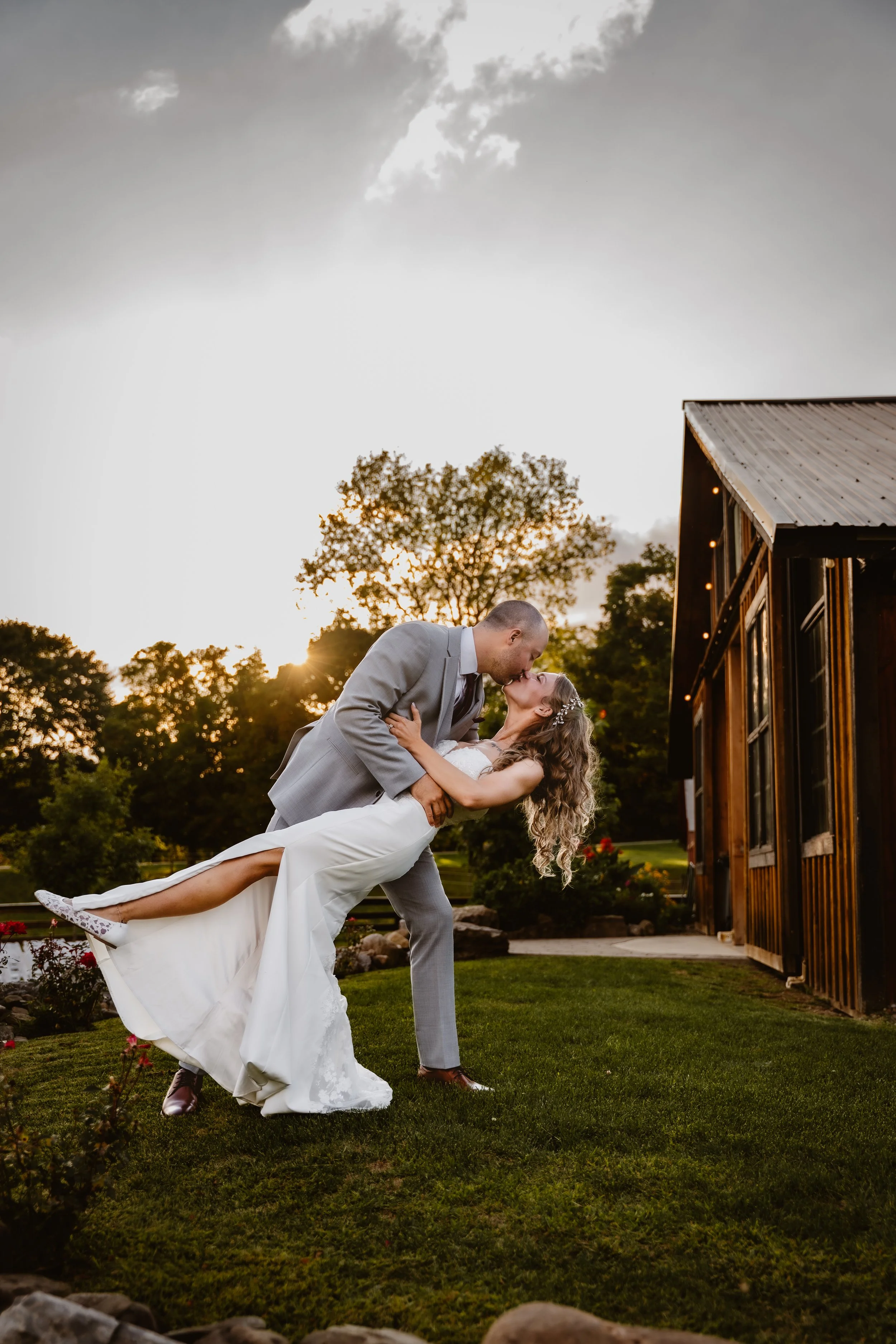 A newlywed couple sharing a kiss outdoors during sunset, with greenery and a wooden building in the background. CHQ Barn, Mayville NY