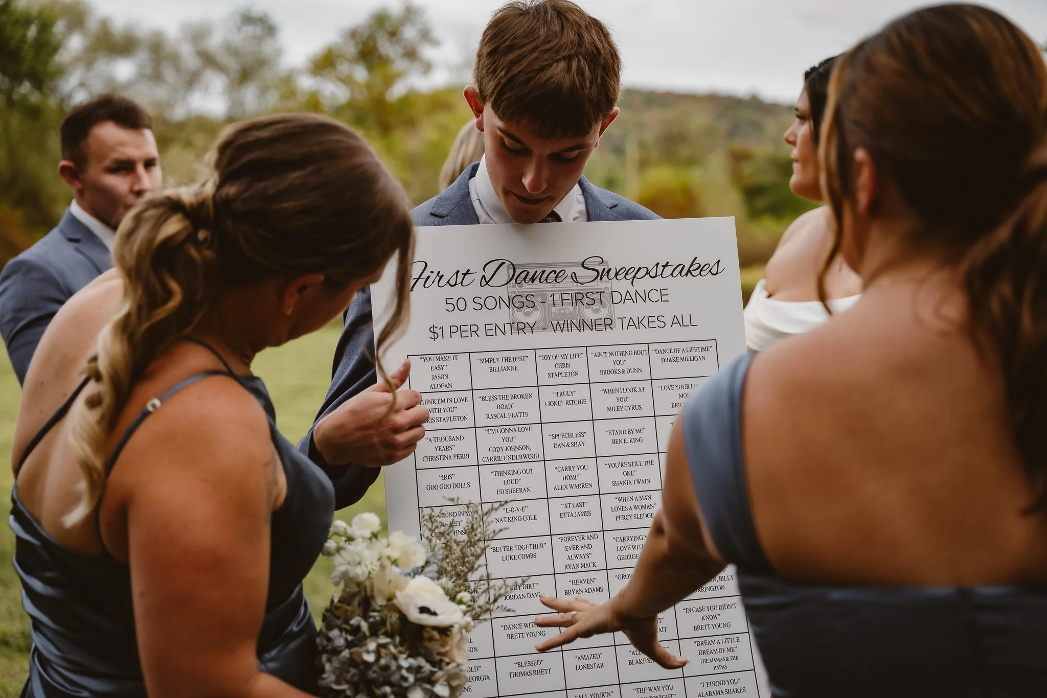Group of people at an outdoor wedding reception looking at a large poster with dance song options for a sweepstakes. Ellicotville Brewing Company, Little Valley, NY. EBC