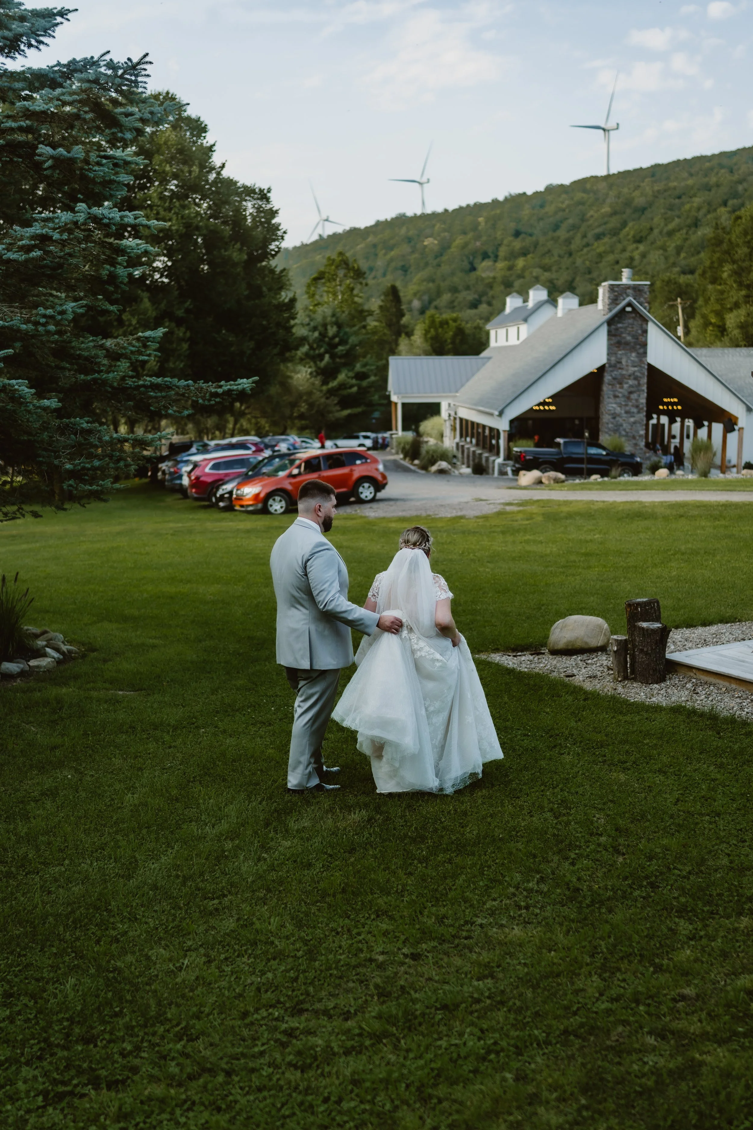 A bride and groom walking outdoors on a grassy area during their wedding, with a building, parked cars, trees, and wind turbines in the background. The Barn at Farrington Hollow, Cherry Creek, NY