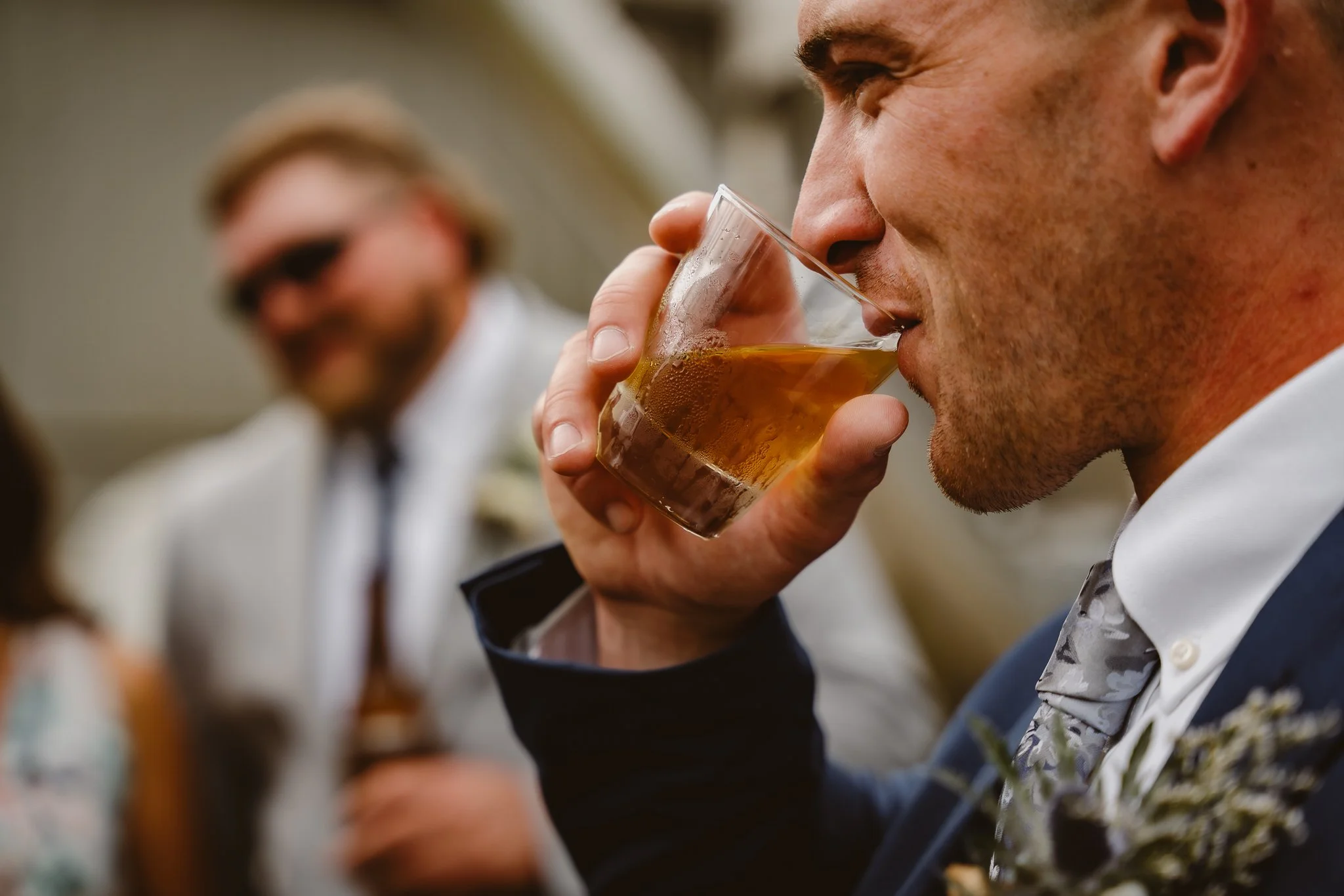 A man in a suit and tie is drinking a beverage from a glass, with a man in the background and a man wearing sunglasses in the background, smiling.