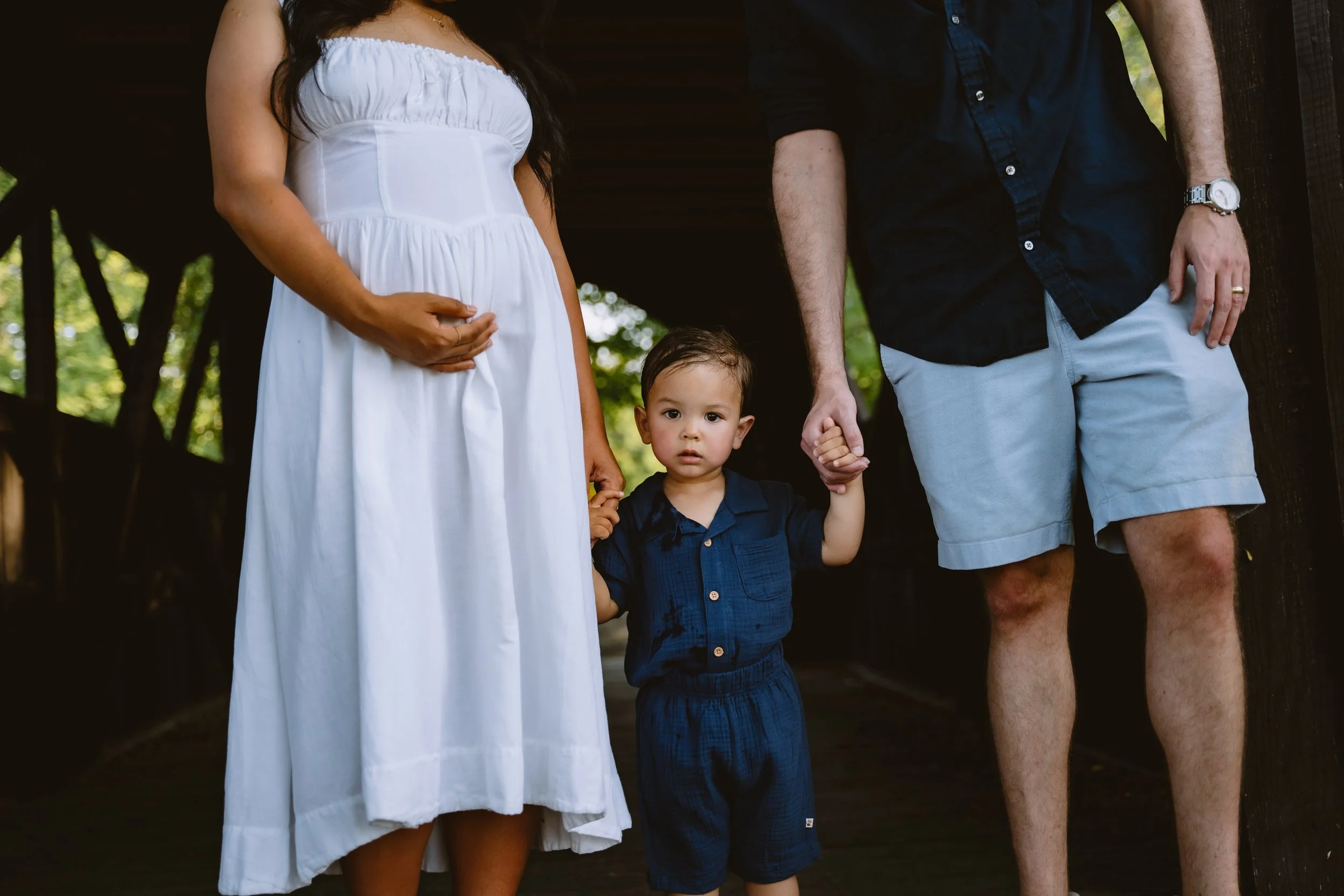 A pregnant woman in a white dress holding her belly, a young boy in a navy blue shirt and shorts holding hands with a man in a dark shirt and light shorts, standing outdoors in a shaded area.