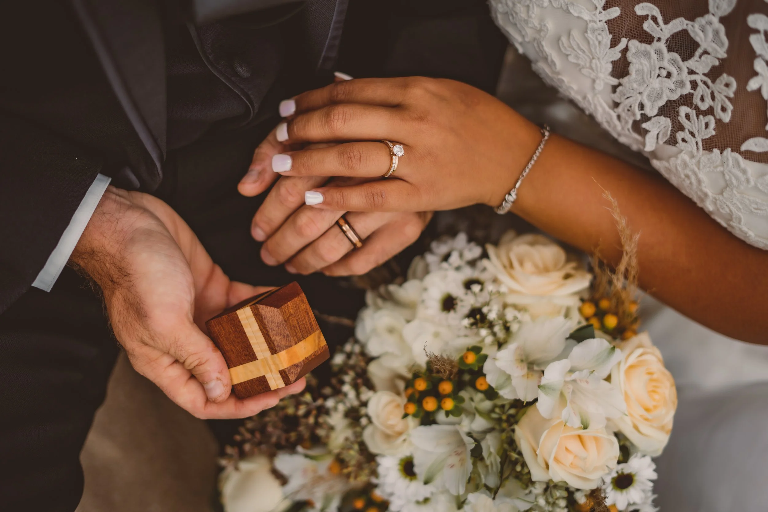 Close-up of a bride and groom holding hands during their wedding ceremony. The bride's hand with an engagement ring and wedding band rests on top of the groom’s hand. The bride is holding a bouquet of white and yellow flowers. The groom is holding a 