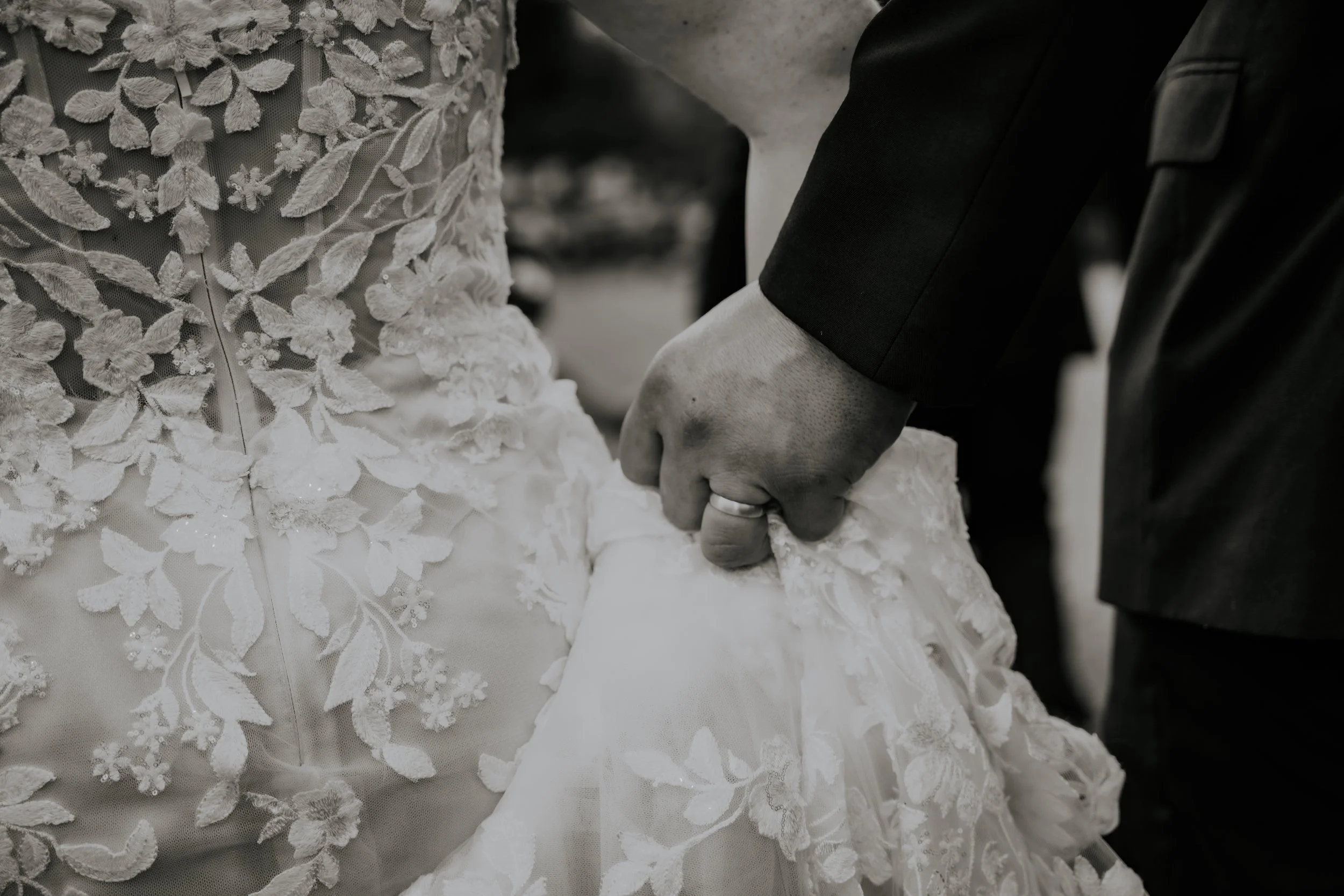 Close-up of a couple holding hands, with the woman wearing an embroidered wedding gown and the man in a dark suit, focusing on their hands and wedding rings.