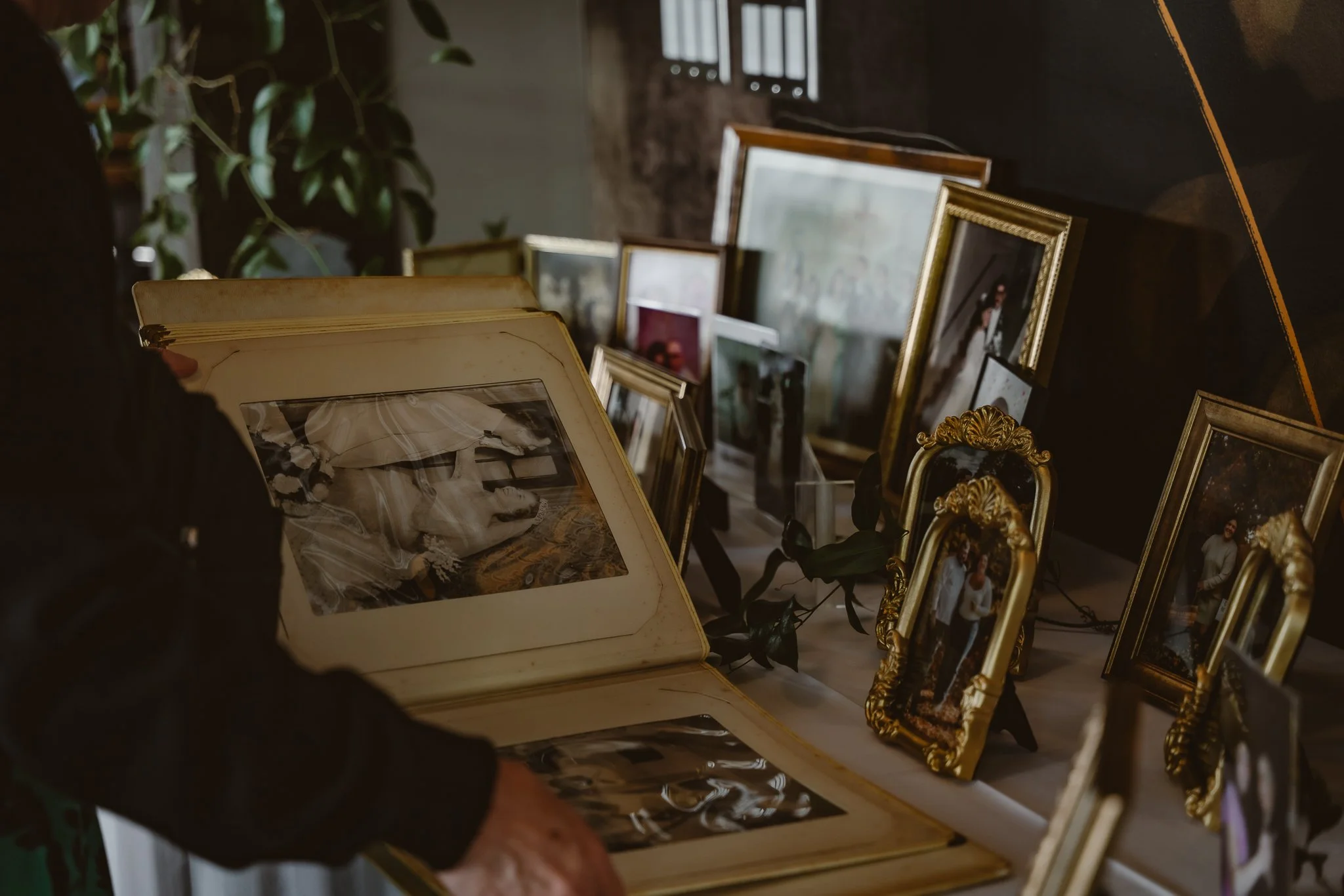 Person holding an open photo album with black and white photographs, alongside various framed photos on a table, some with ornate gold frames, in a dimly lit room.