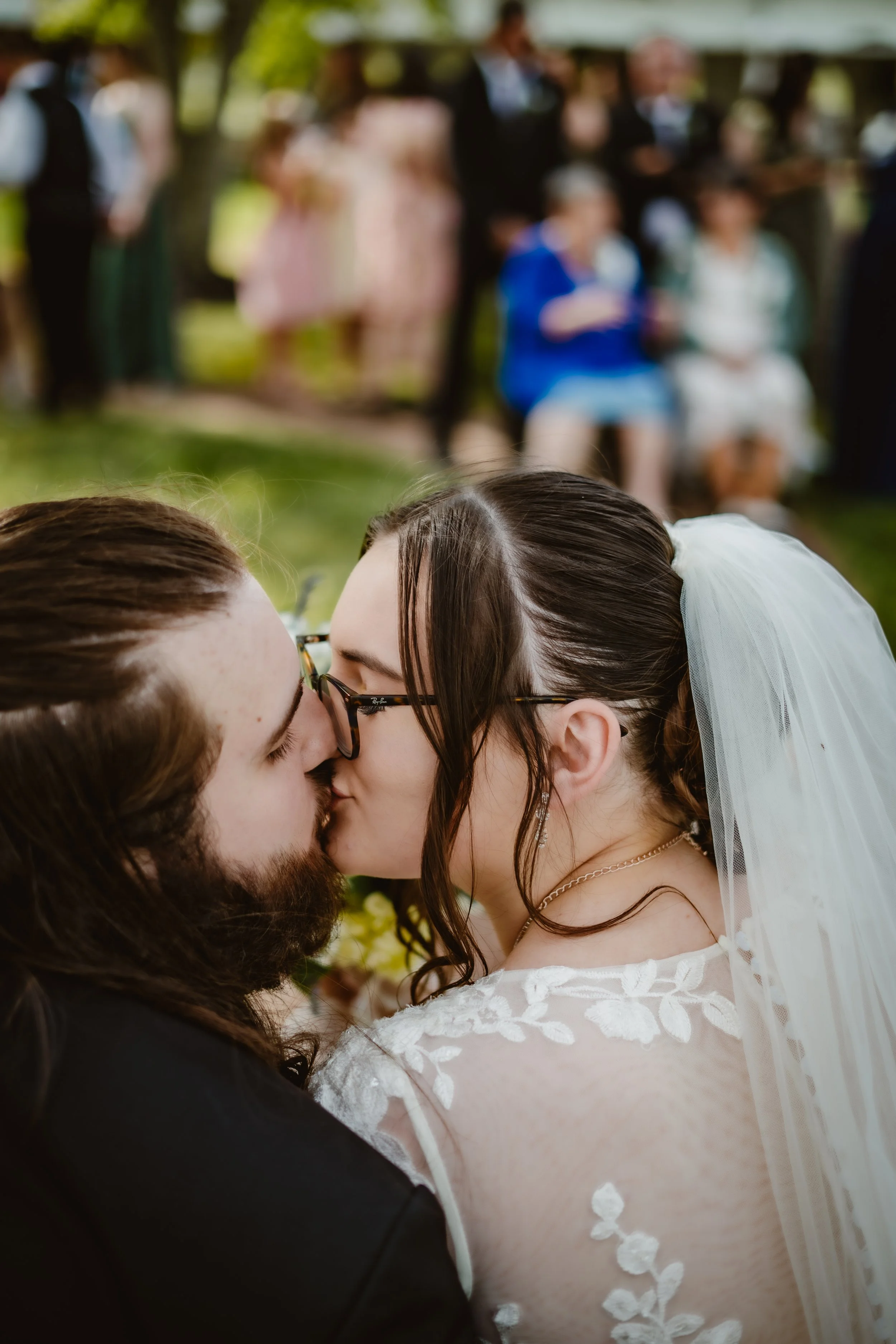 A bride and groom kiss during their wedding ceremony outdoors, with guests blurred in the background.