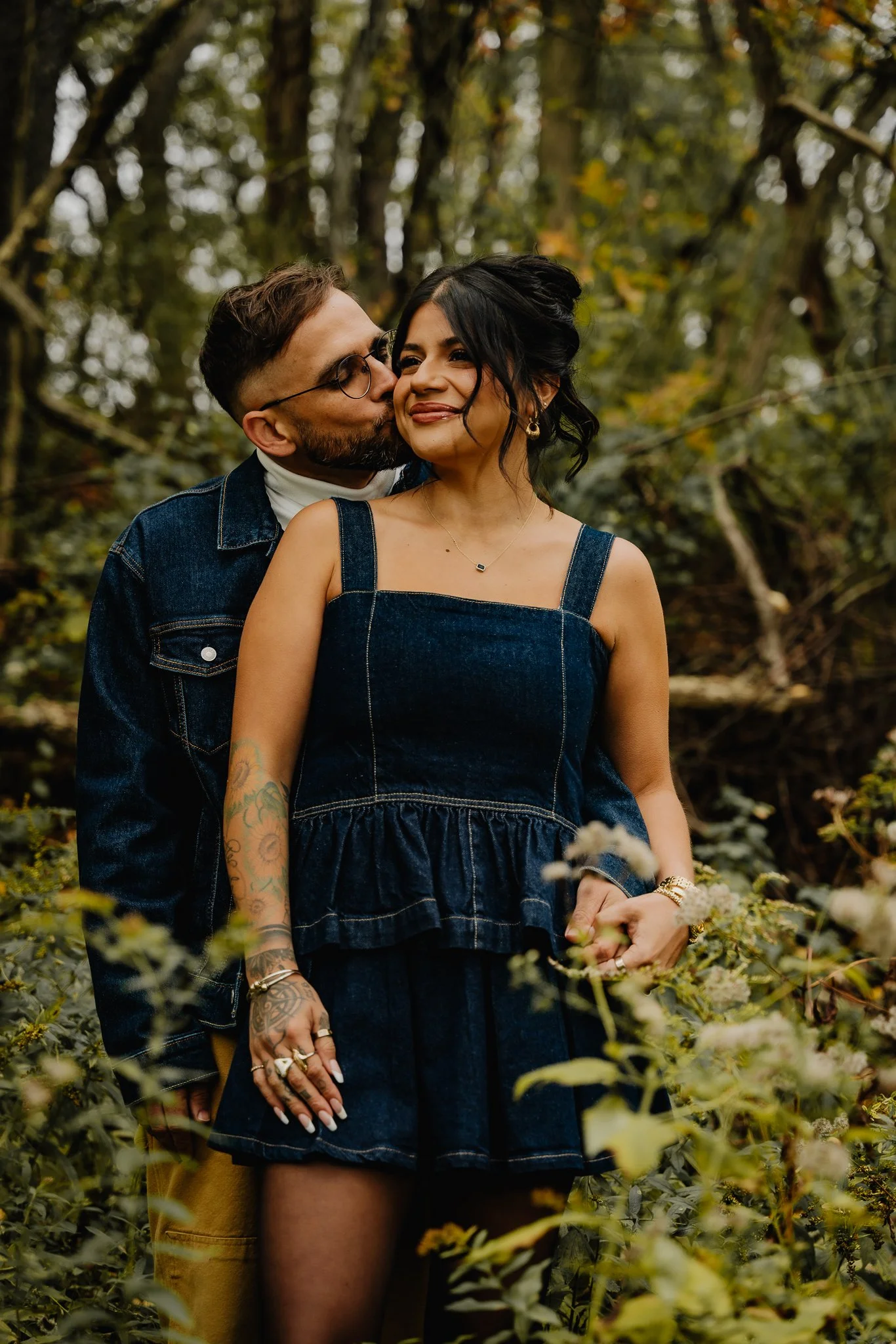 A couple in denim outfits standing close together outdoors among green foliage, with the man kissing the woman's cheek and the woman smiling.
