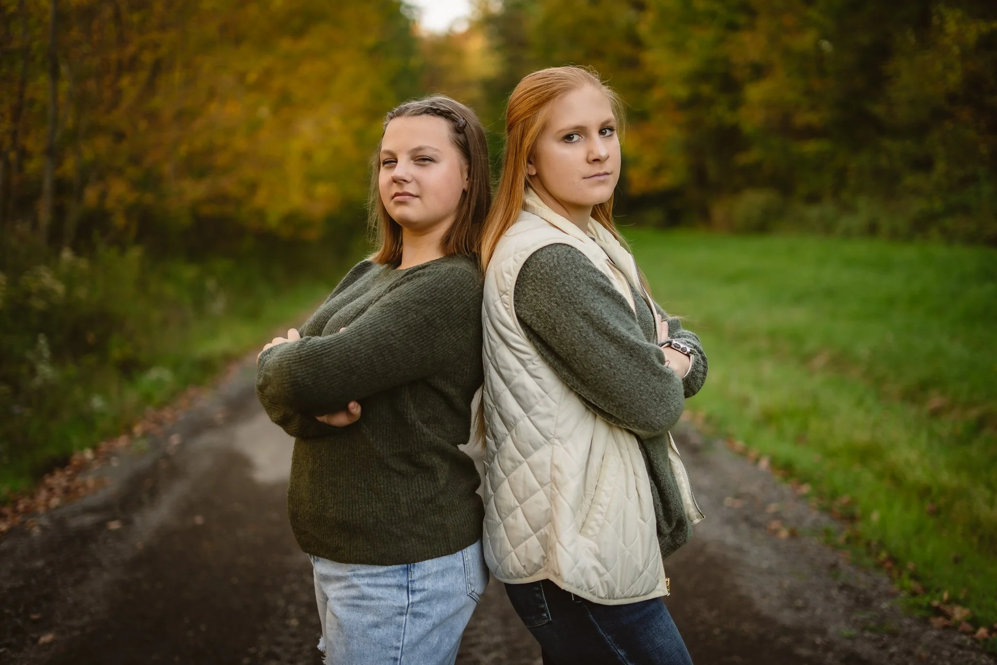 Two young women stand back to back with crossed arms on a fall outdoor trail.