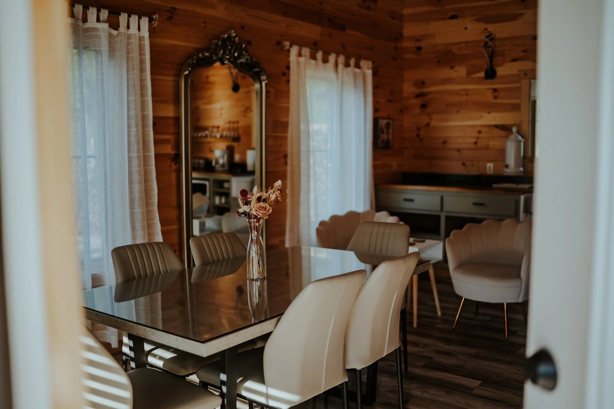 Dining room with a wooden wall, a glass-top table with a floral centerpiece, beige chairs, and curtains on windows. Farrington Hollow, Cherry Creek, NY