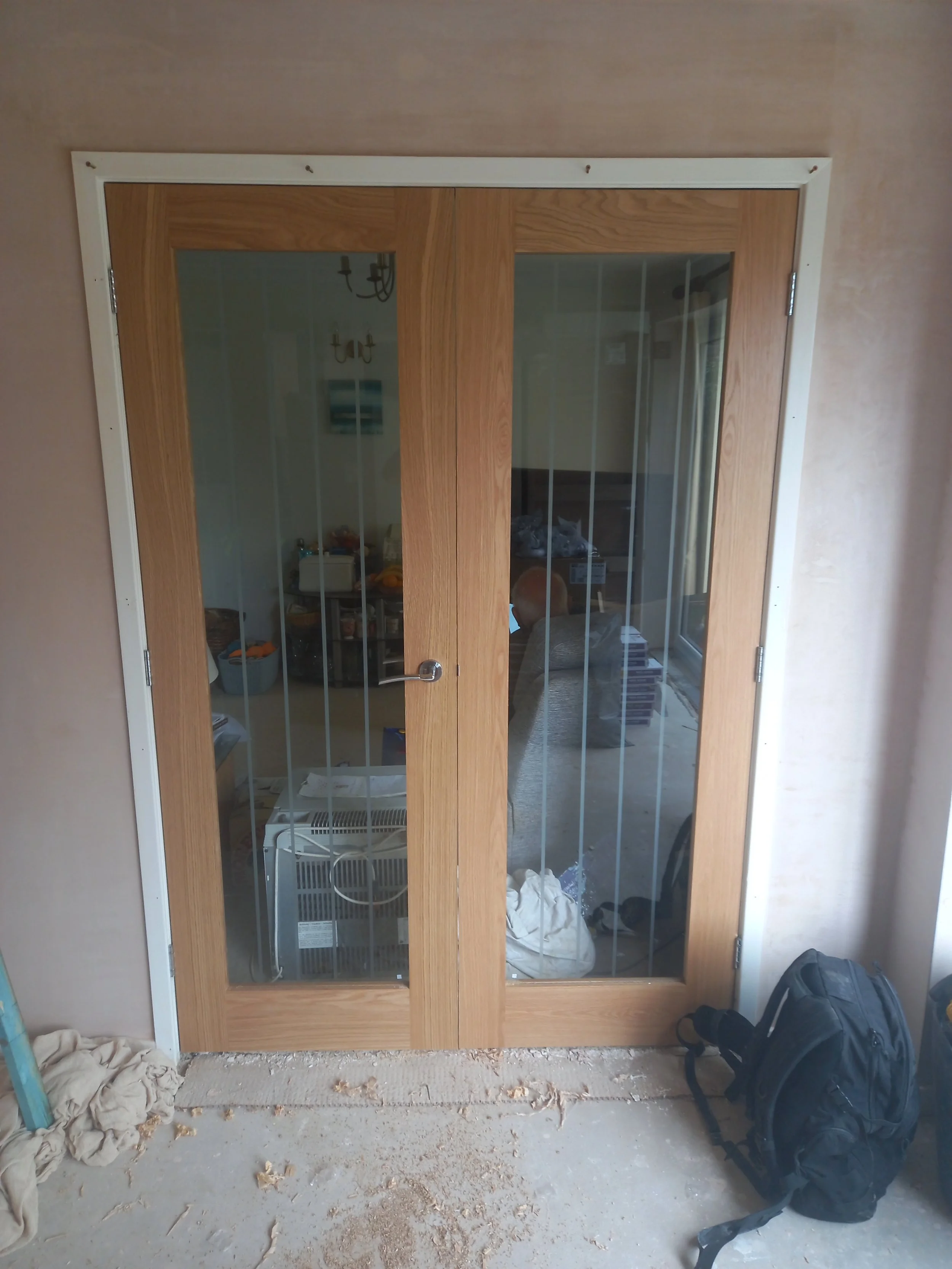 Wooden framed glass double doors with black metal handles set in a beige wall, leading into a room with furniture and artwork, with construction debris and a black backpack on the floor in the foreground.