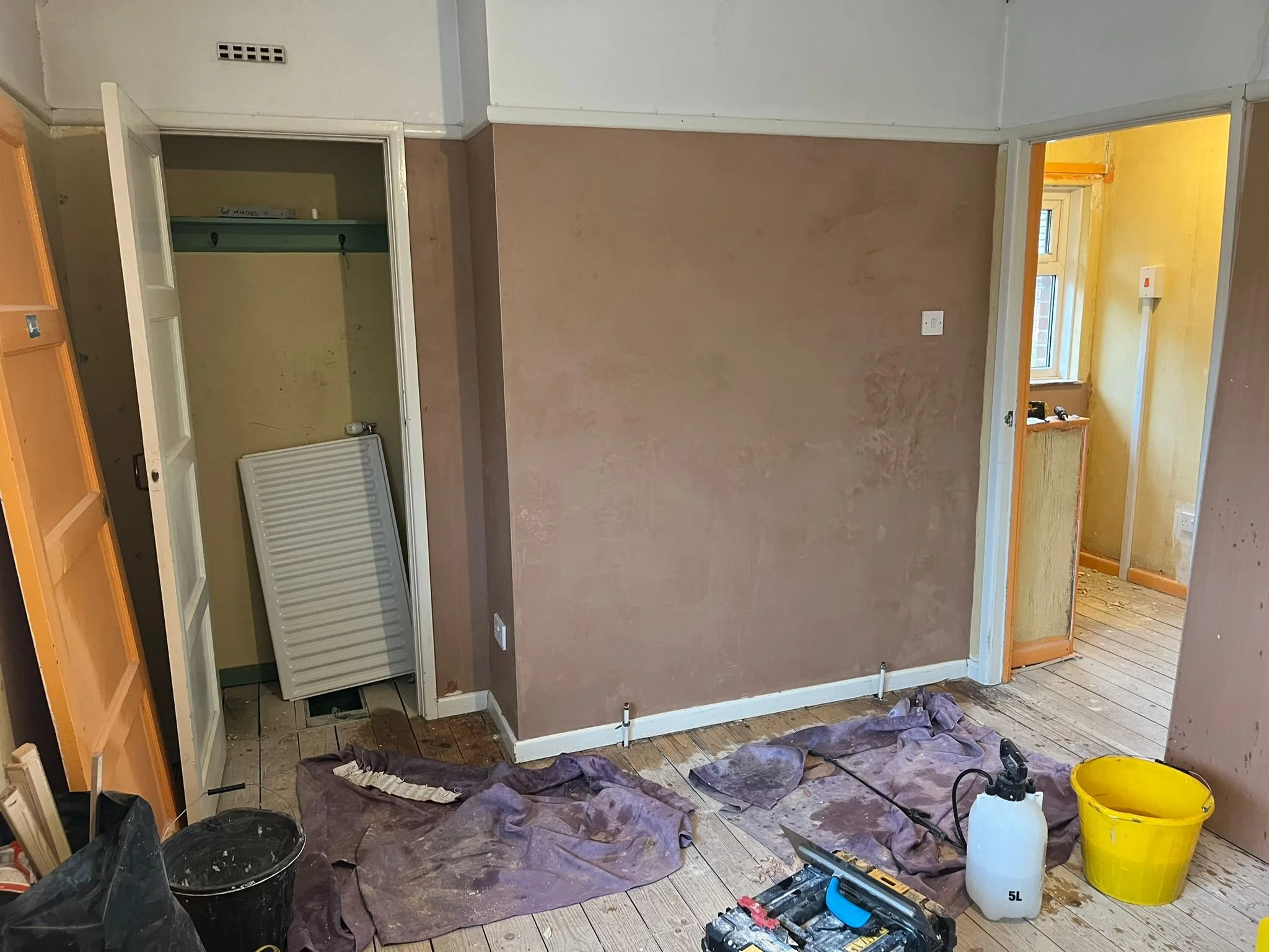 A room under renovation with partially painted walls, construction tools, and cleaning supplies scattered on the floor, including paint buckets, a spray bottle, and tarps.