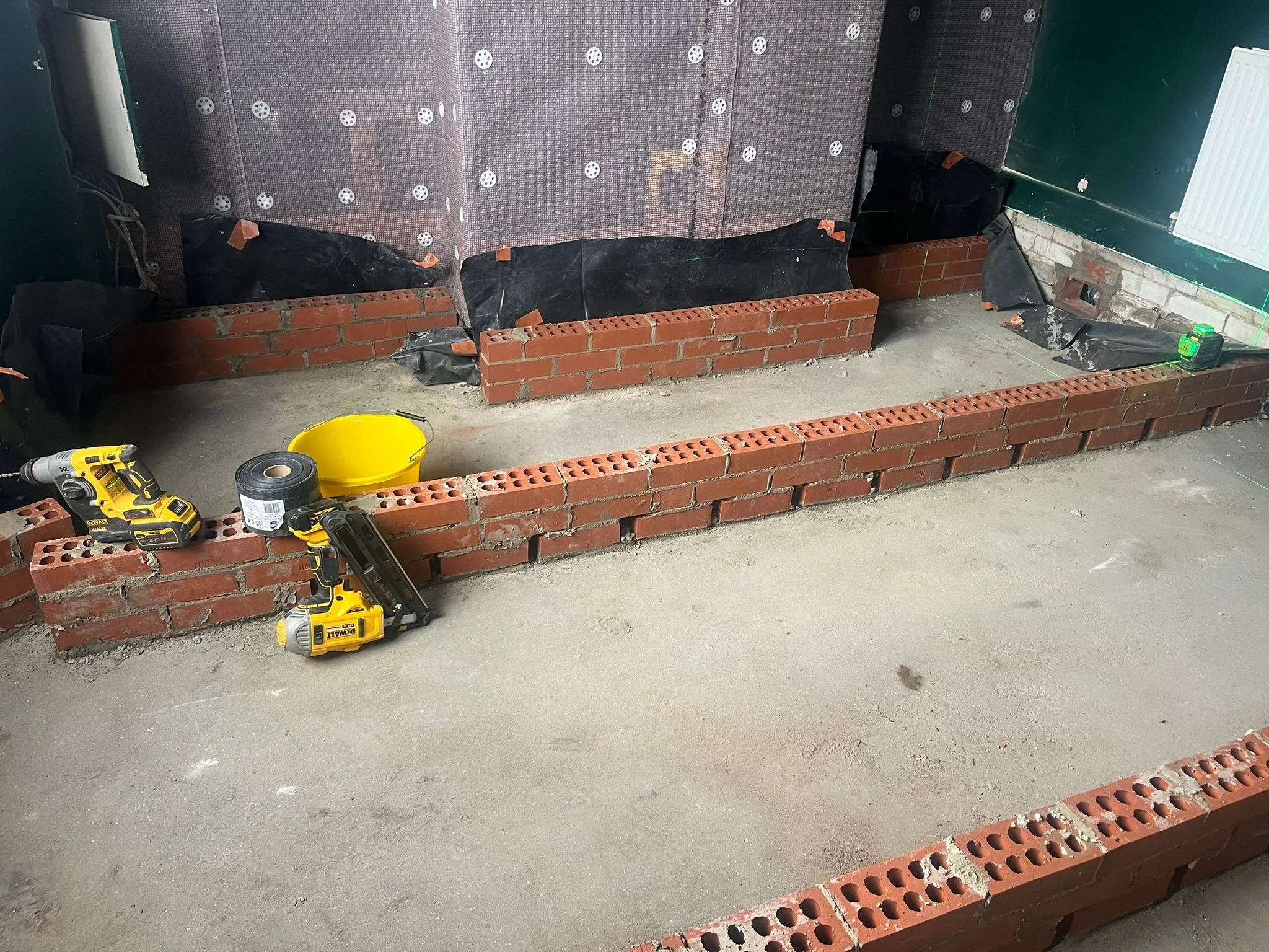 Construction site with a partially built brick wall, construction tools, and supplies including a cordless drill, tape measure, black plastic sheeting, and a bucket, with a concrete floor and a green door in the background.