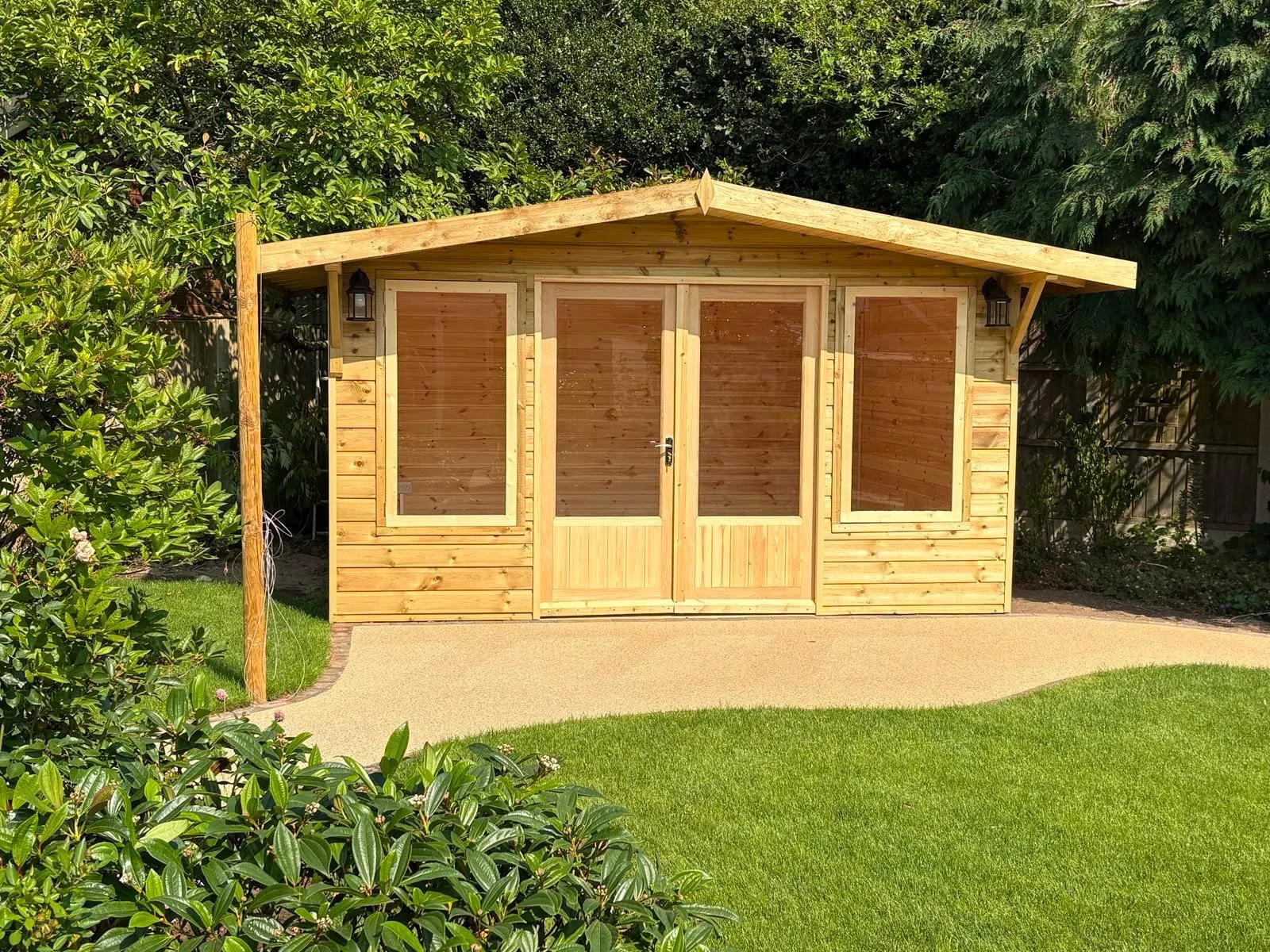 A small wooden garden shed with double doors and three large windows, situated on a concrete pathway in a backyard with green grass and shrubbery.
