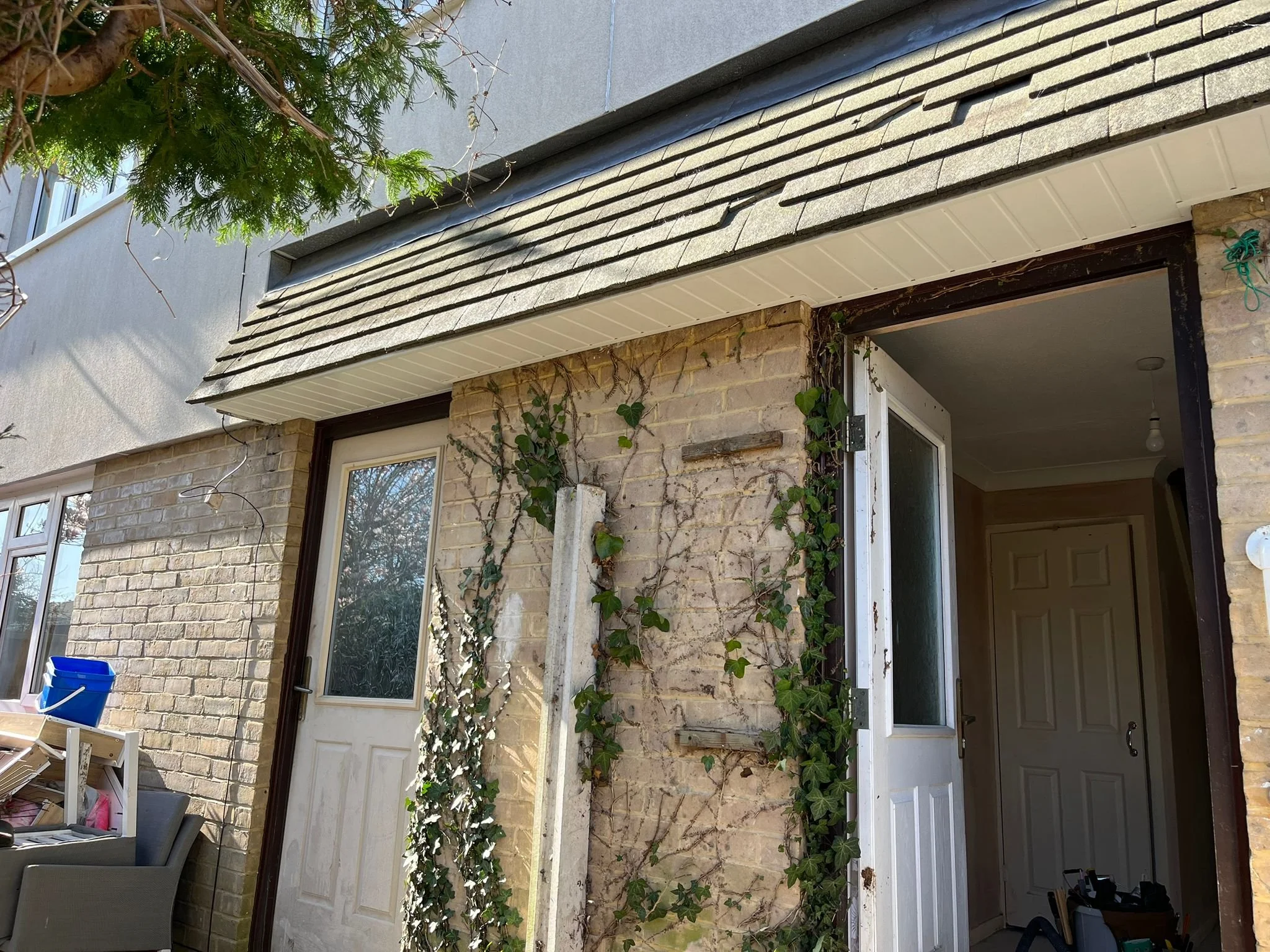 The back of a house showing a brick wall and vinyl siding, with a door opening into an interior room. A vine climbs along the brick wall. Part of a roof with shingles is visible, and a tree with green foliage is partly in frame.