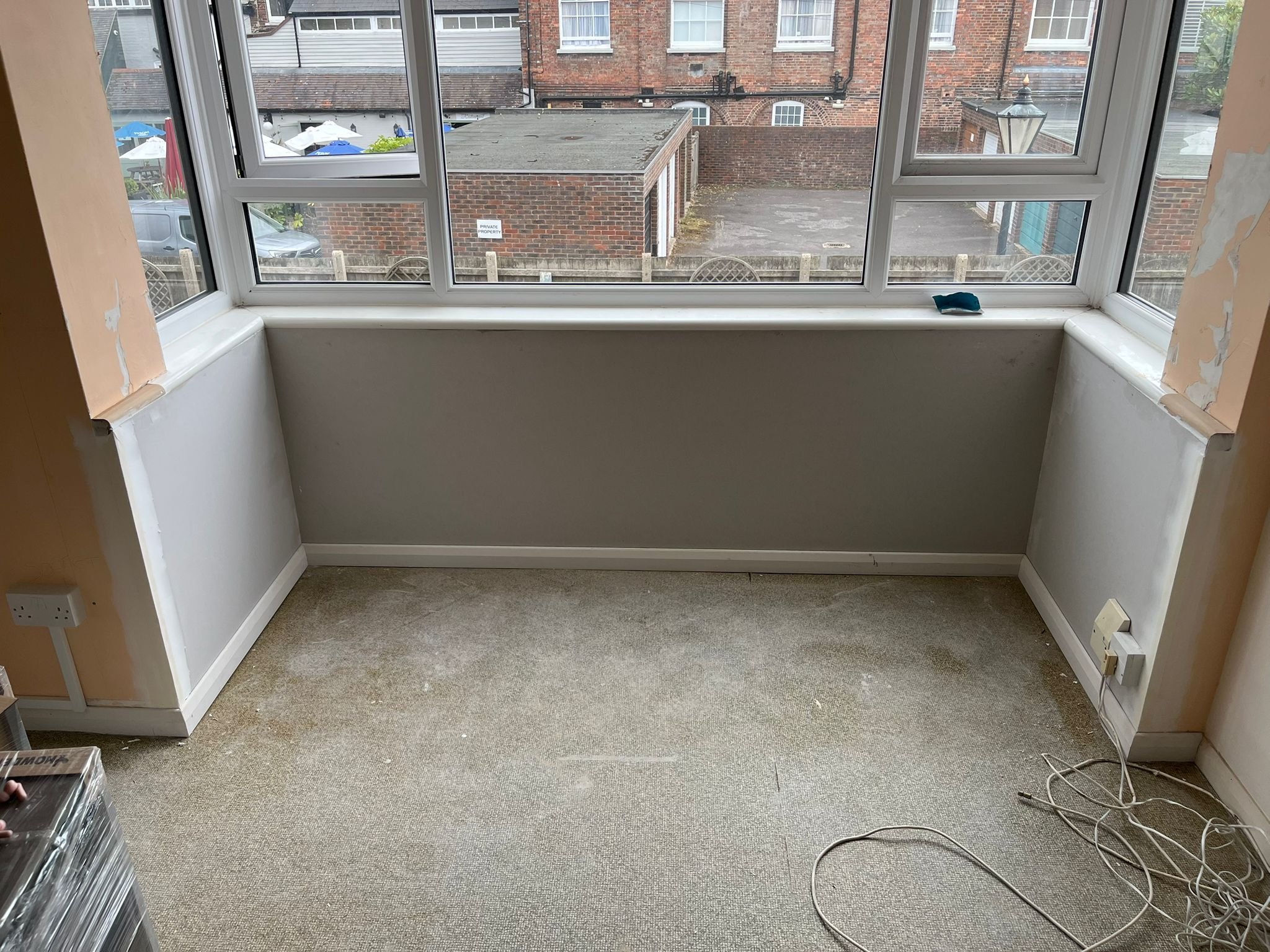 Unfinished room with beige carpet, torn walls, and a large window overlooking a brick building and paved area outside.