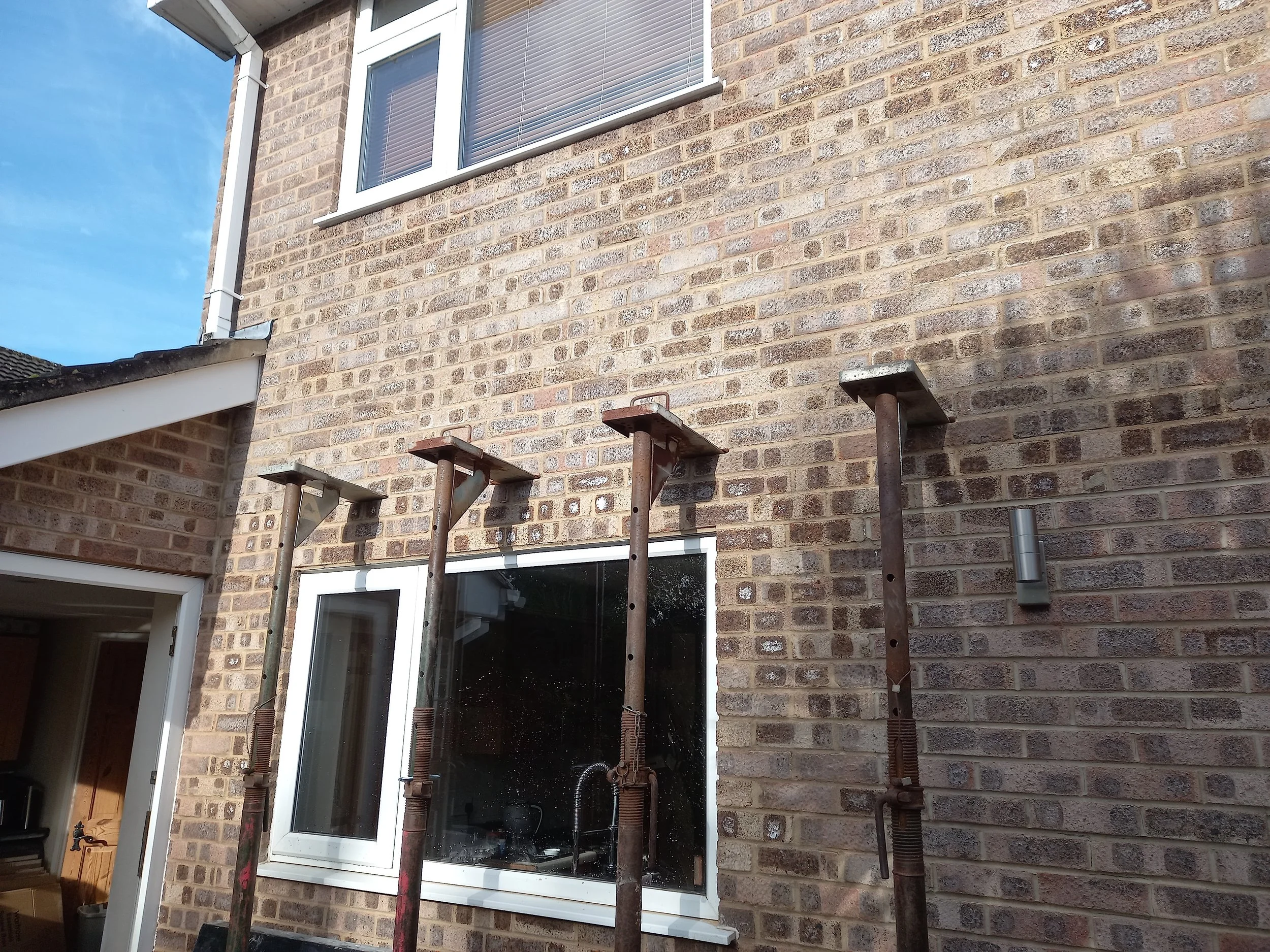Close-up of a brick wall exterior with two large windows and scaffolding supports in front, some of which are rusty.