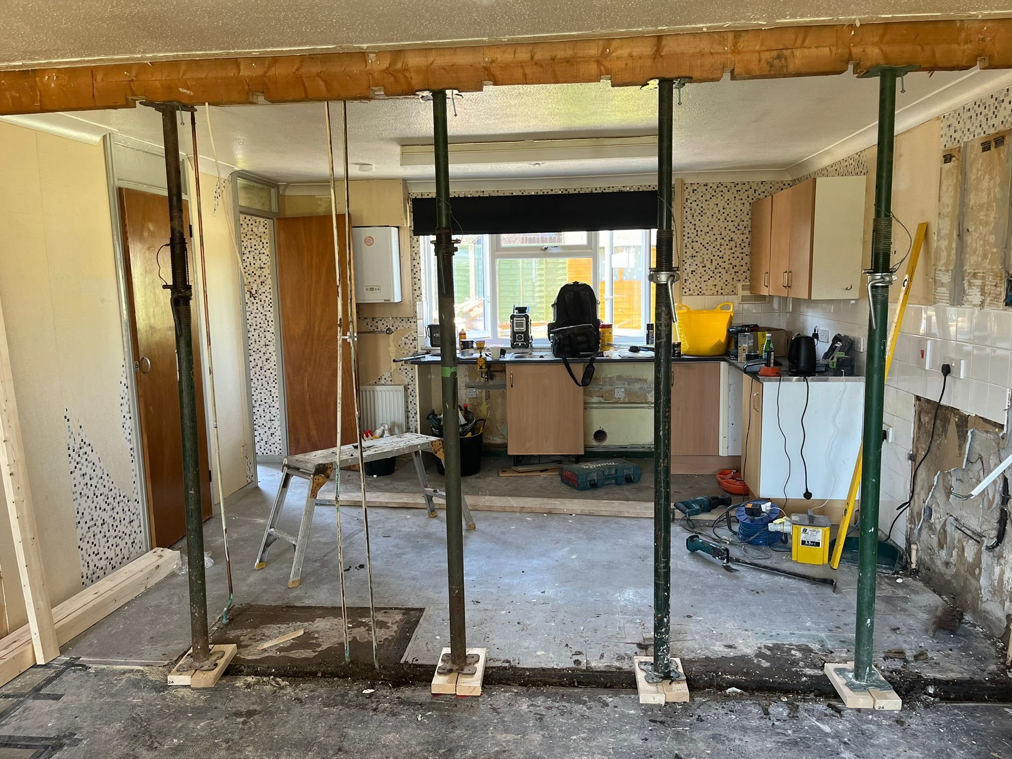 Kitchen under renovation with scaffolding, tools, workbench, yellow bucket, and exposed wall sections.