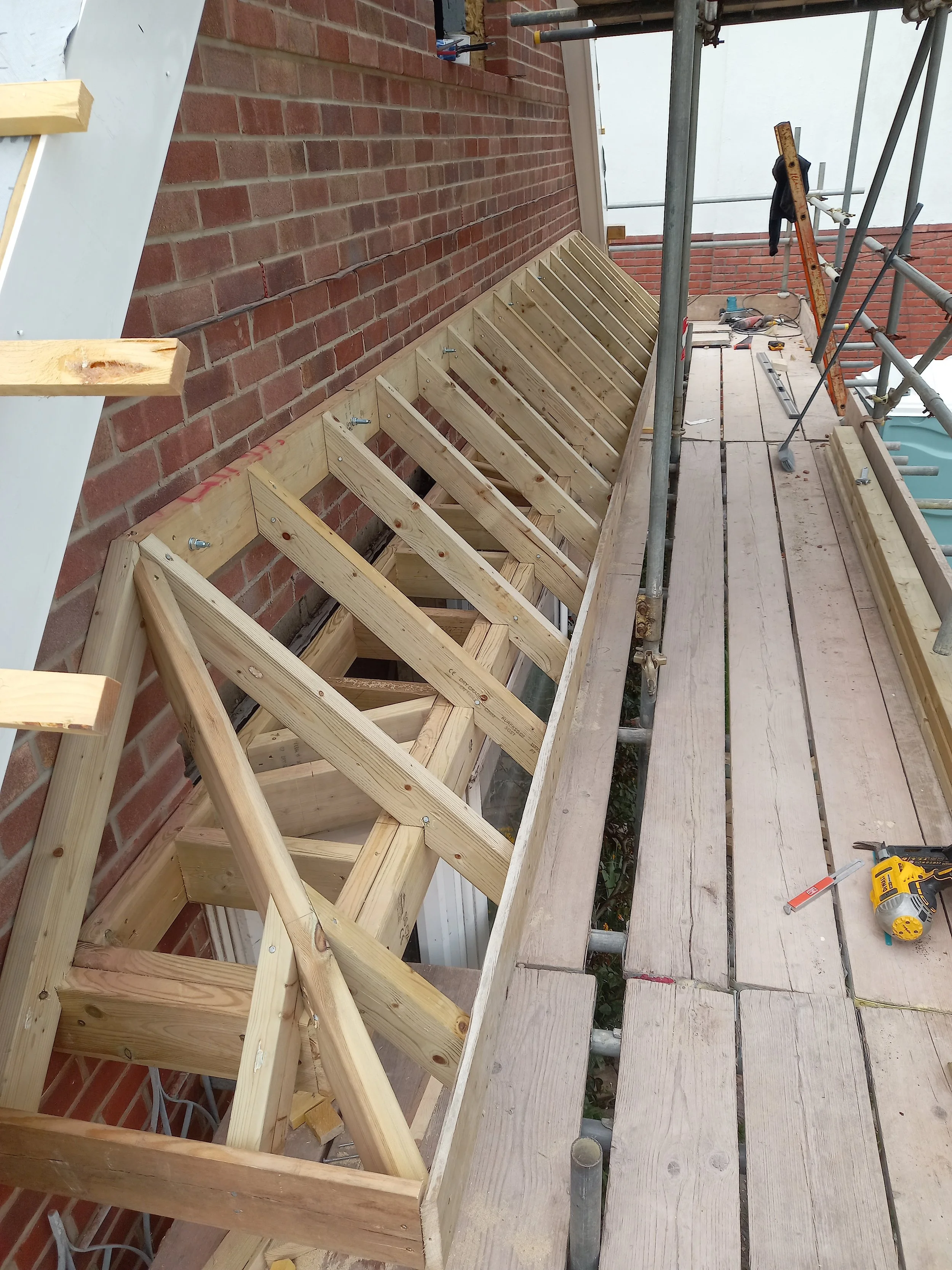 Construction site with wooden framework and scaffolding adjacent to a brick wall, tools and materials scattered on the planked floor.