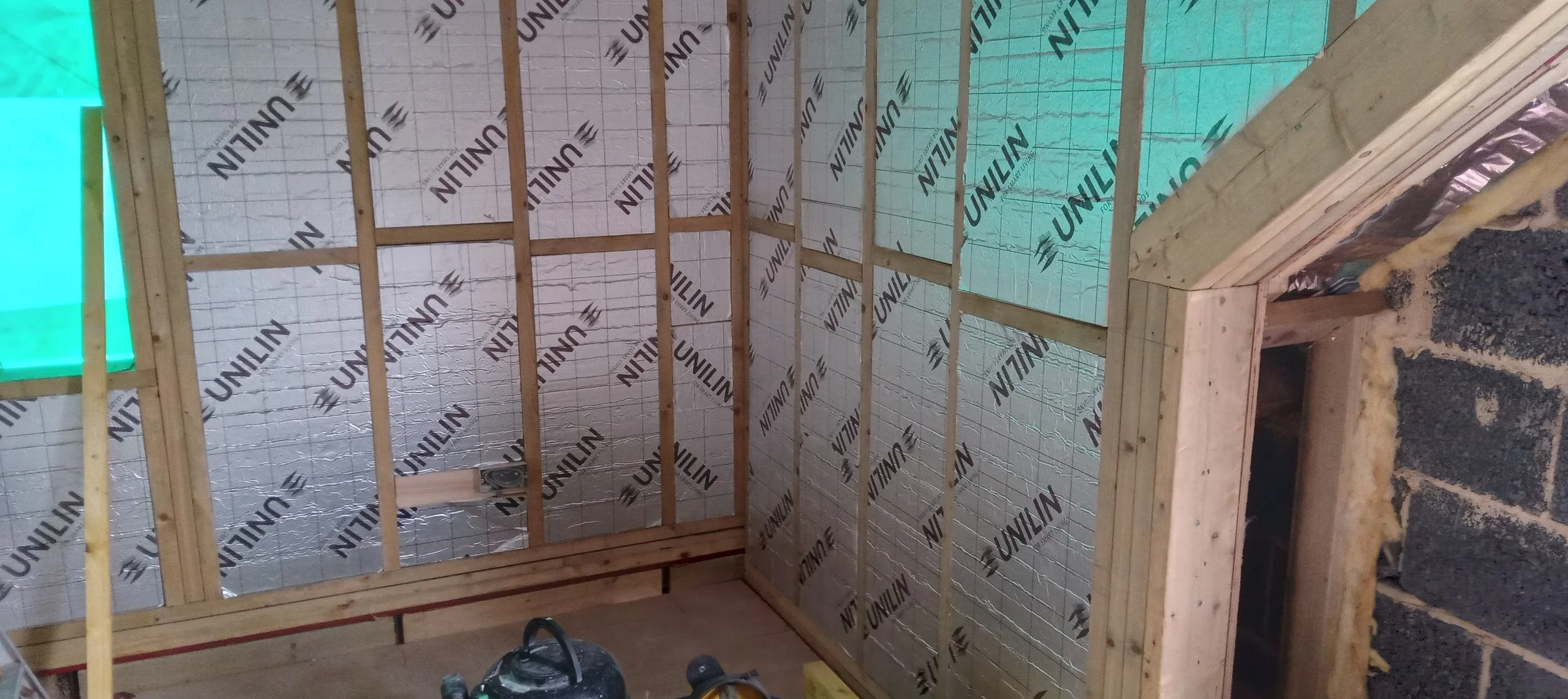 Interior view of a wooden-framed room under construction, with insulation installed on the walls and ceiling, and a small opening on the right side.