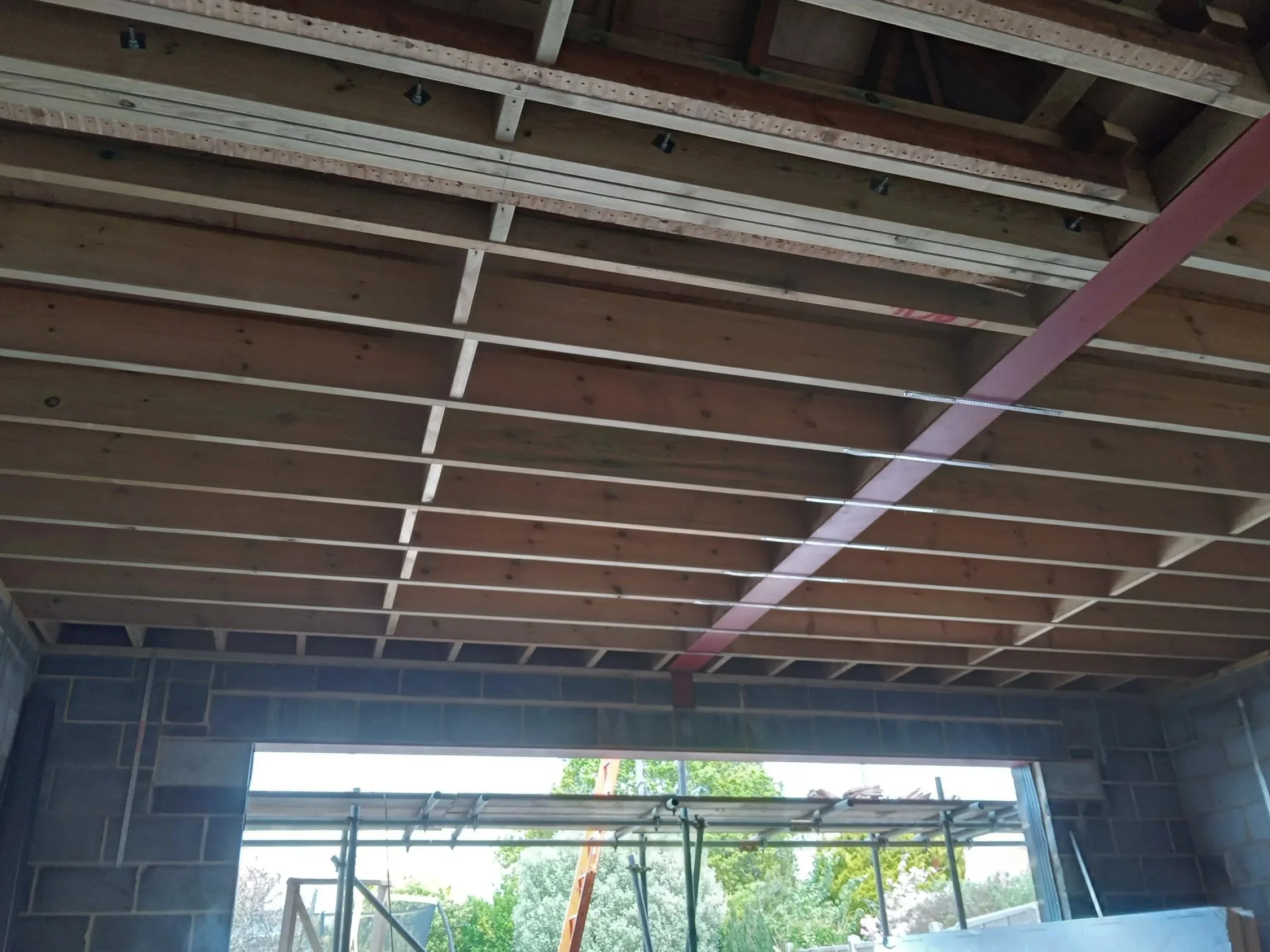 The image shows the wooden framework of a ceiling under construction, with exposed beams and scaffolding visible outside through a large open window.
