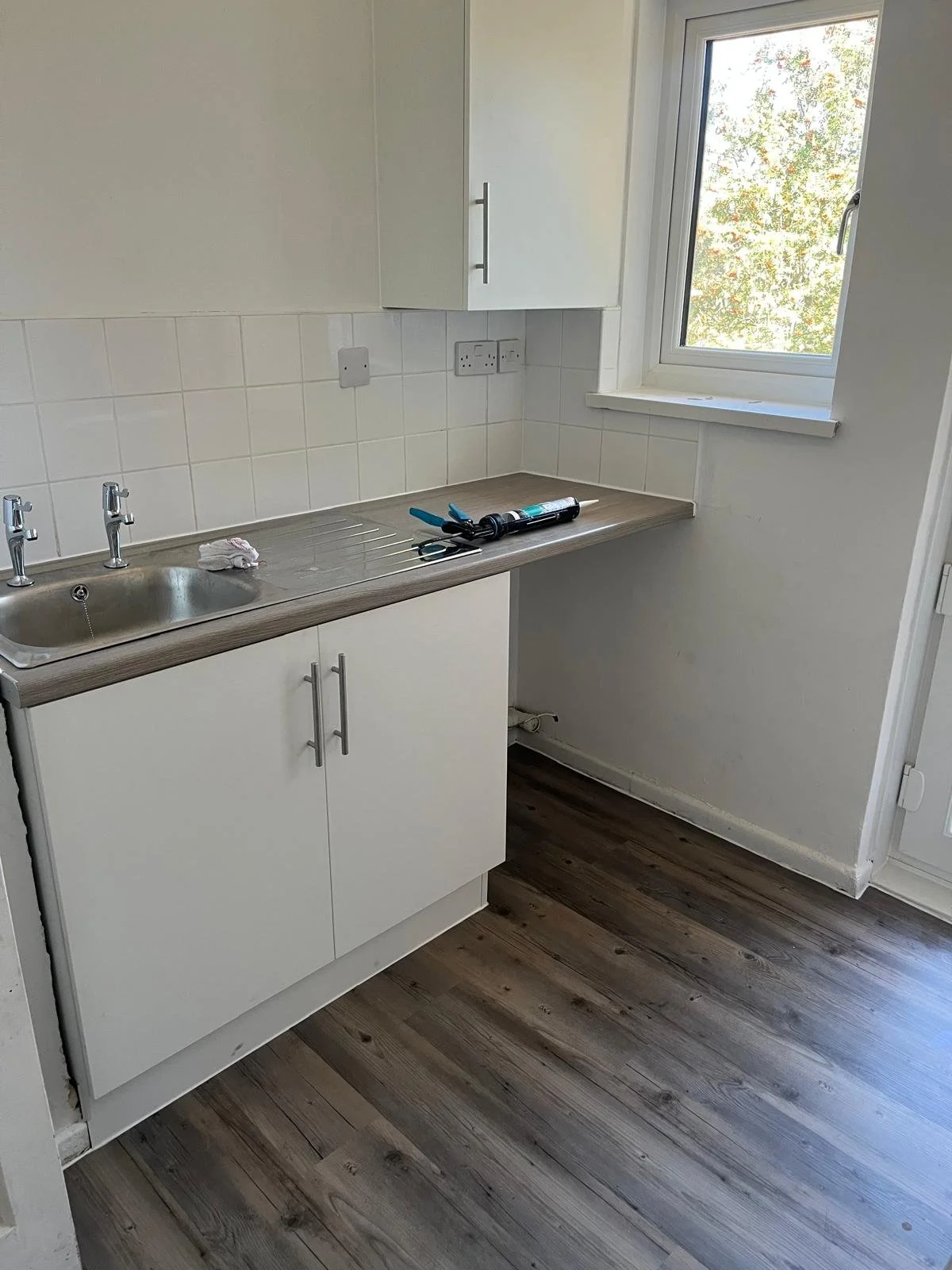 A small kitchen with a white cabinet and countertop, a stainless steel sink, a window with trees outside, and some tools and supplies on the counter.