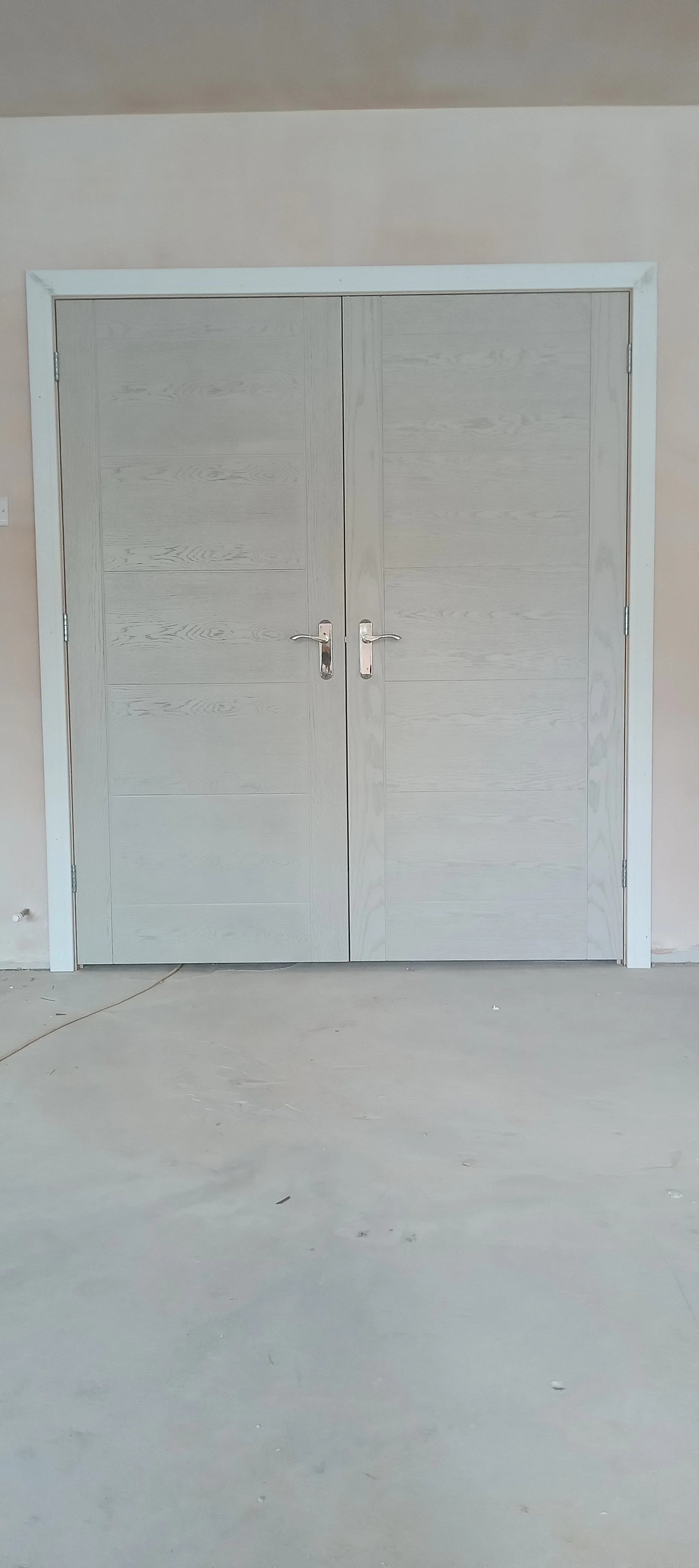 Double gray wooden doors with silver handles set in a beige wall, with a concrete floor in front.