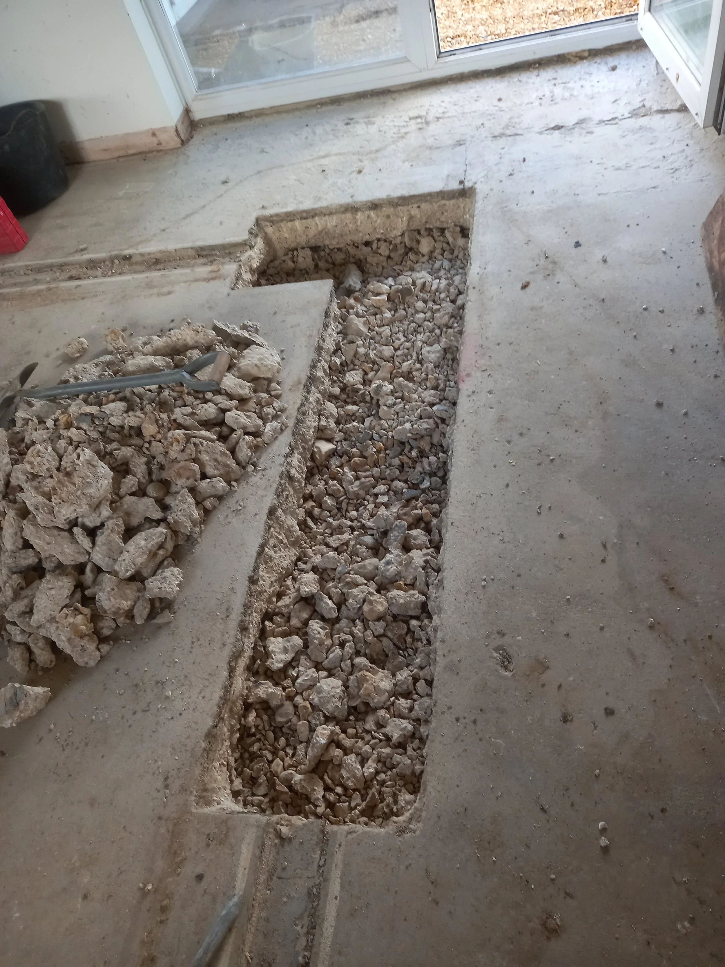 A section of concrete floor removed, exposing dirt and rocks underneath, with construction tools visible on the rubble, next to a sliding glass door.