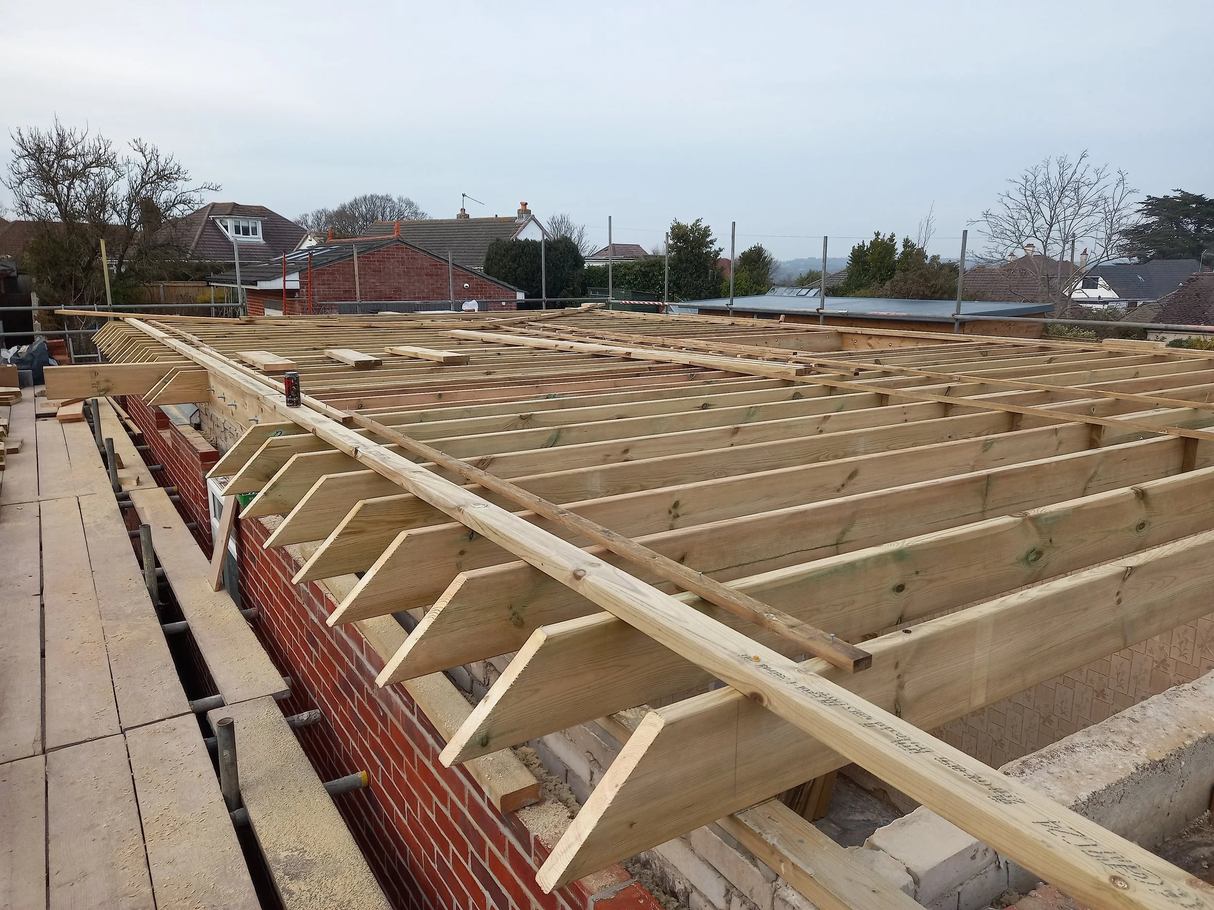 Construction site with wooden roof framing on top of a brick building, with residential houses and trees in the background.