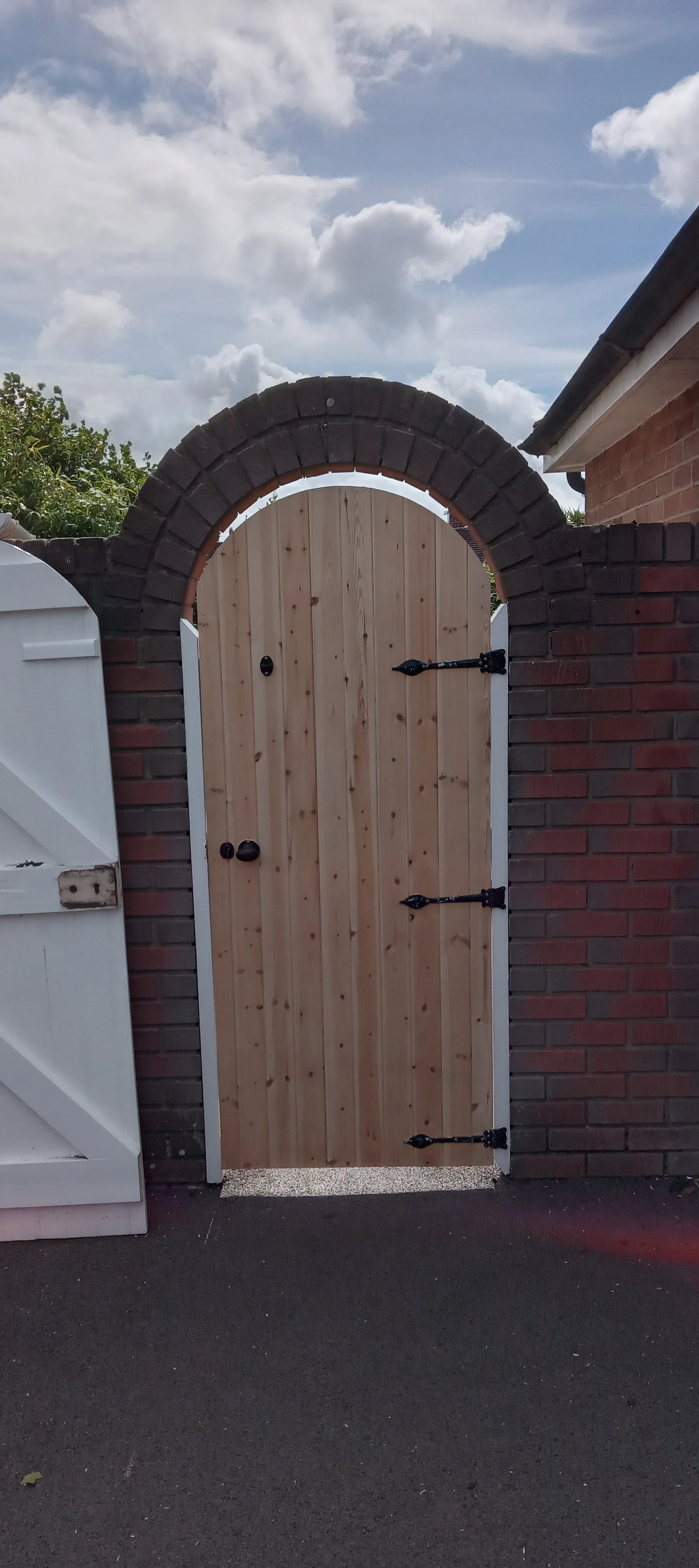 A wooden foot gate with black hinges and latch set into a brick archway, next to a white fence post, under a partly cloudy sky.