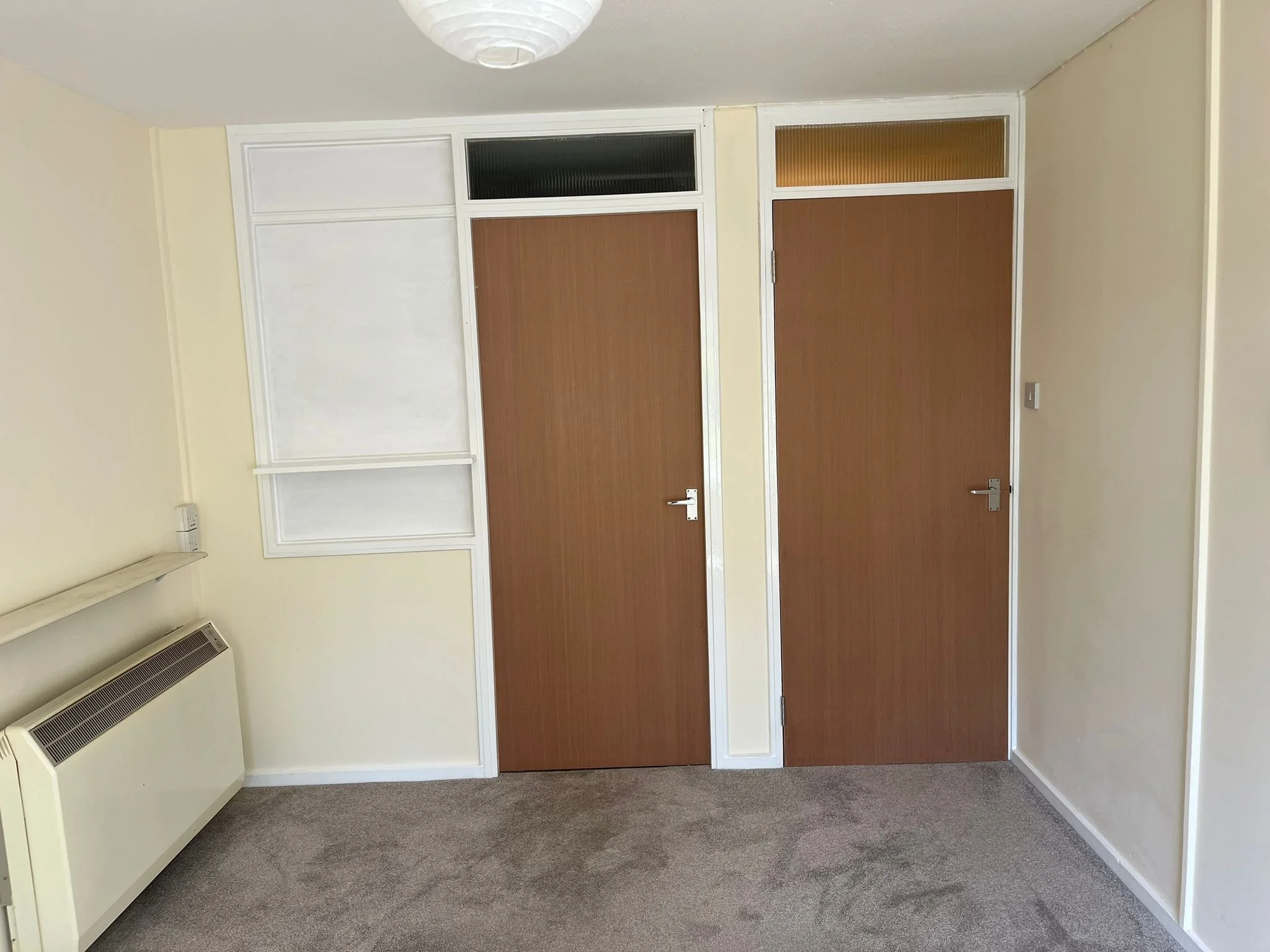 Interior of a room with cream-colored walls, gray carpeted floor, wooden doors with white frames, and a white ceiling-mounted light fixture.