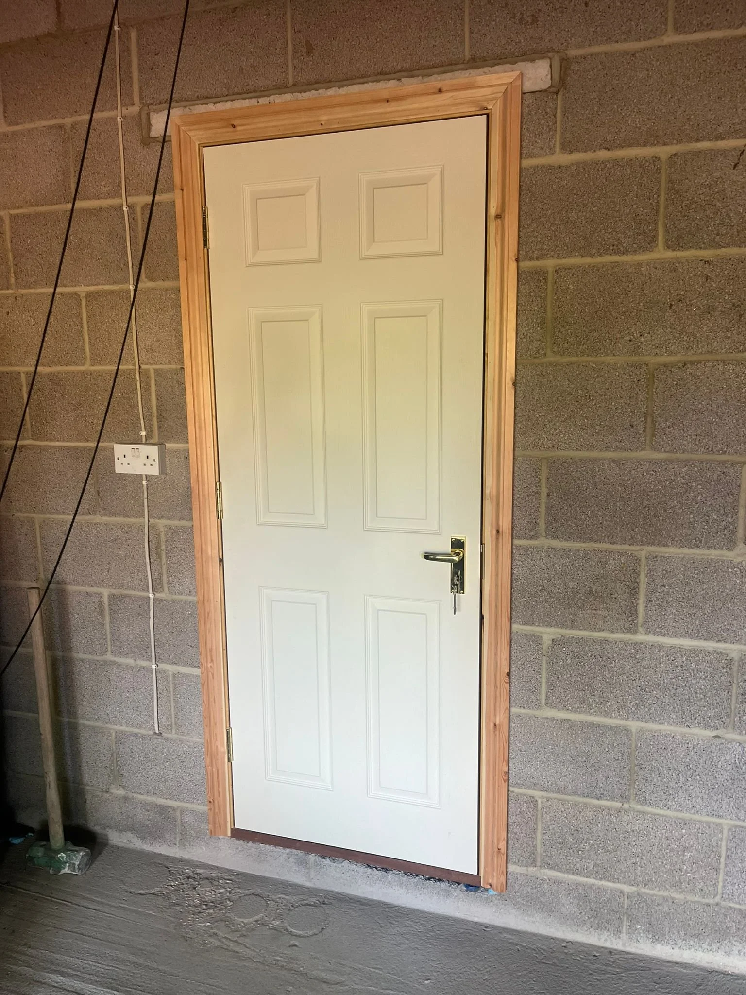 A white door with a gold handle and hinges, framed with unpainted wooden trim, set in a gray cinder block wall inside a construction site or unfinished room.