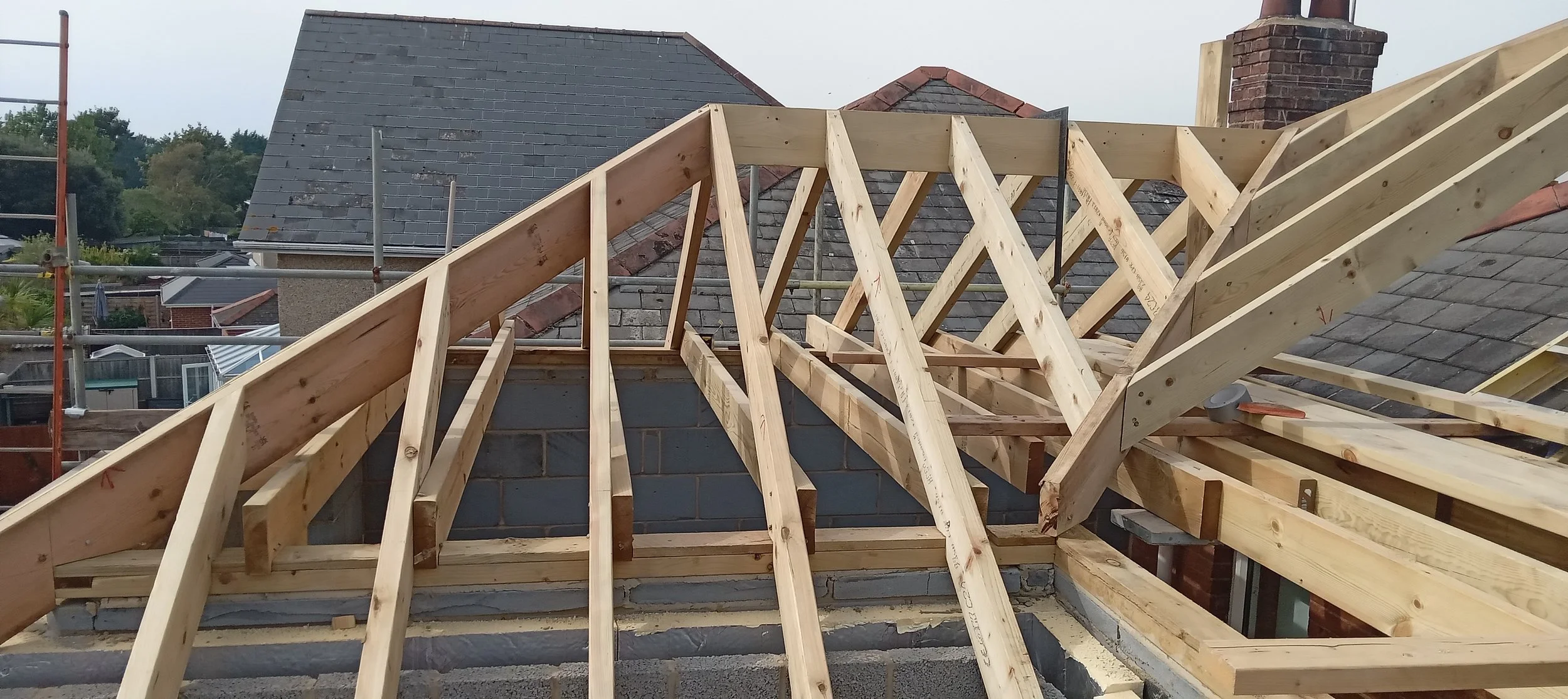 Wooden framework construction for a roof on a building under construction, with scaffolding nearby and neighboring houses visible in the background.
