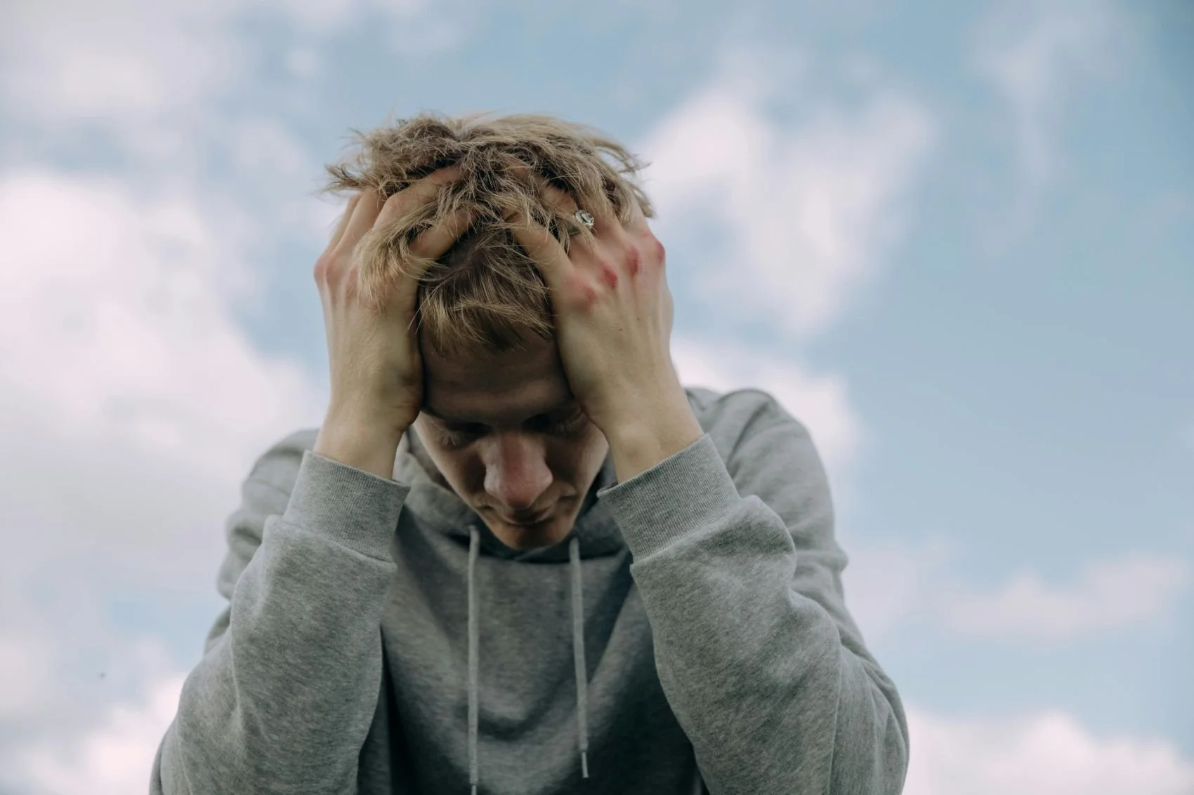 A young man with light brown hair, wearing a gray hoodie, holding his head with both hands, looking down, standing outdoors against a cloudy sky.