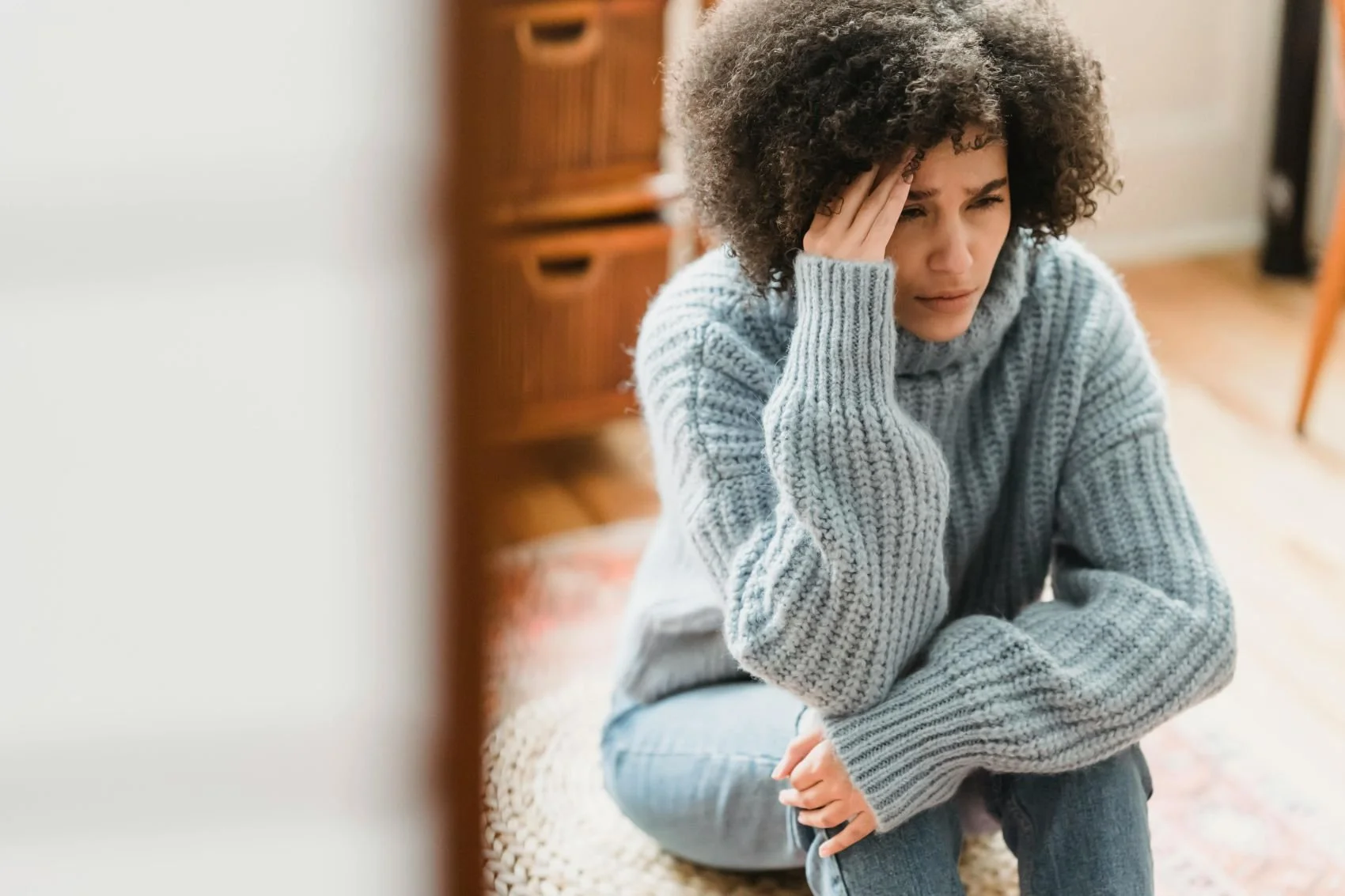 A woman sitting on the floor with a distressed expression, holding her head with one hand, wearing a gray sweater and jeans, in a cozy room.
