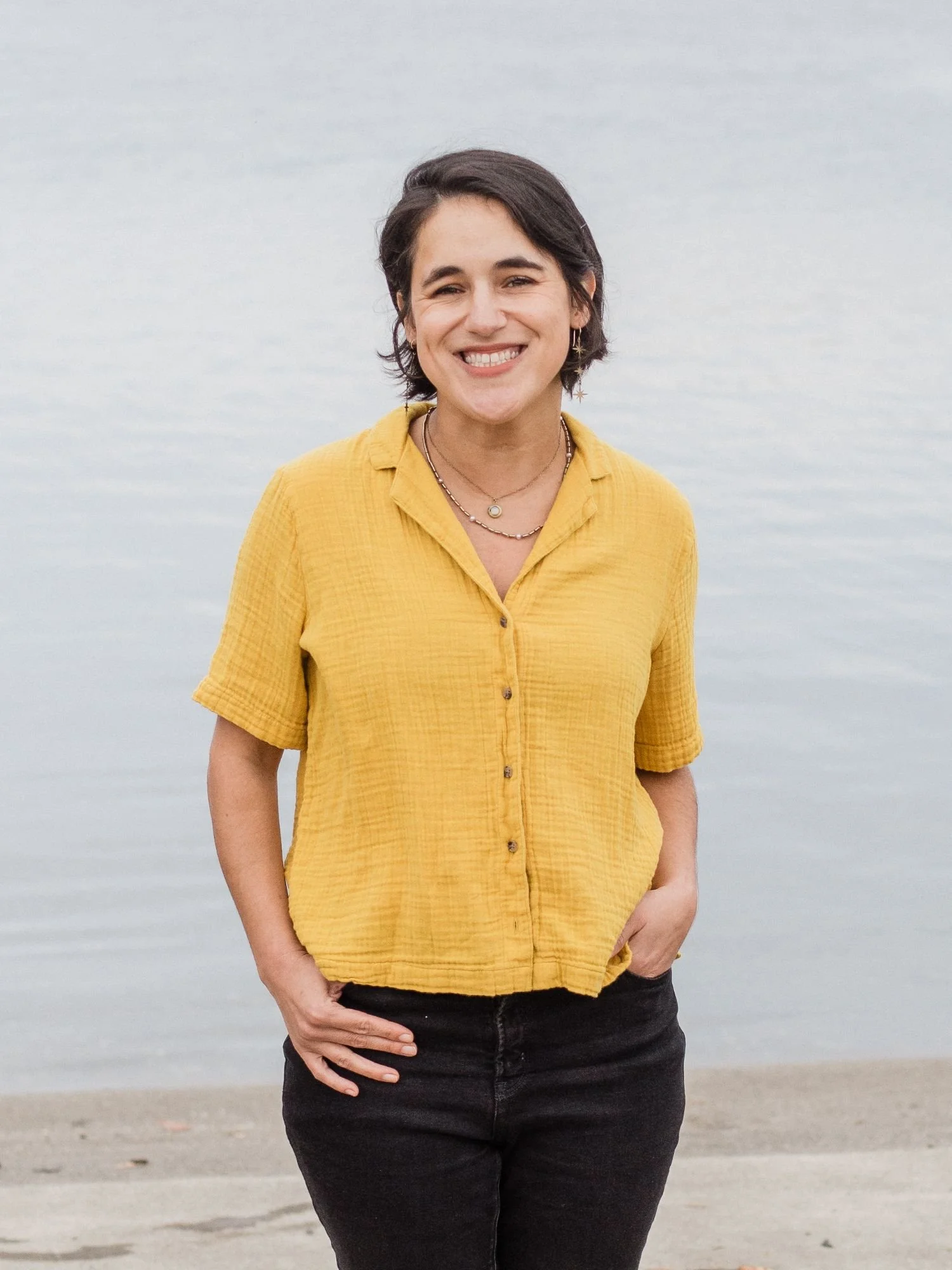 A woman with short dark hair, smiling, wearing a yellow button-up shirt and black pants, standing outdoors near a body of water.