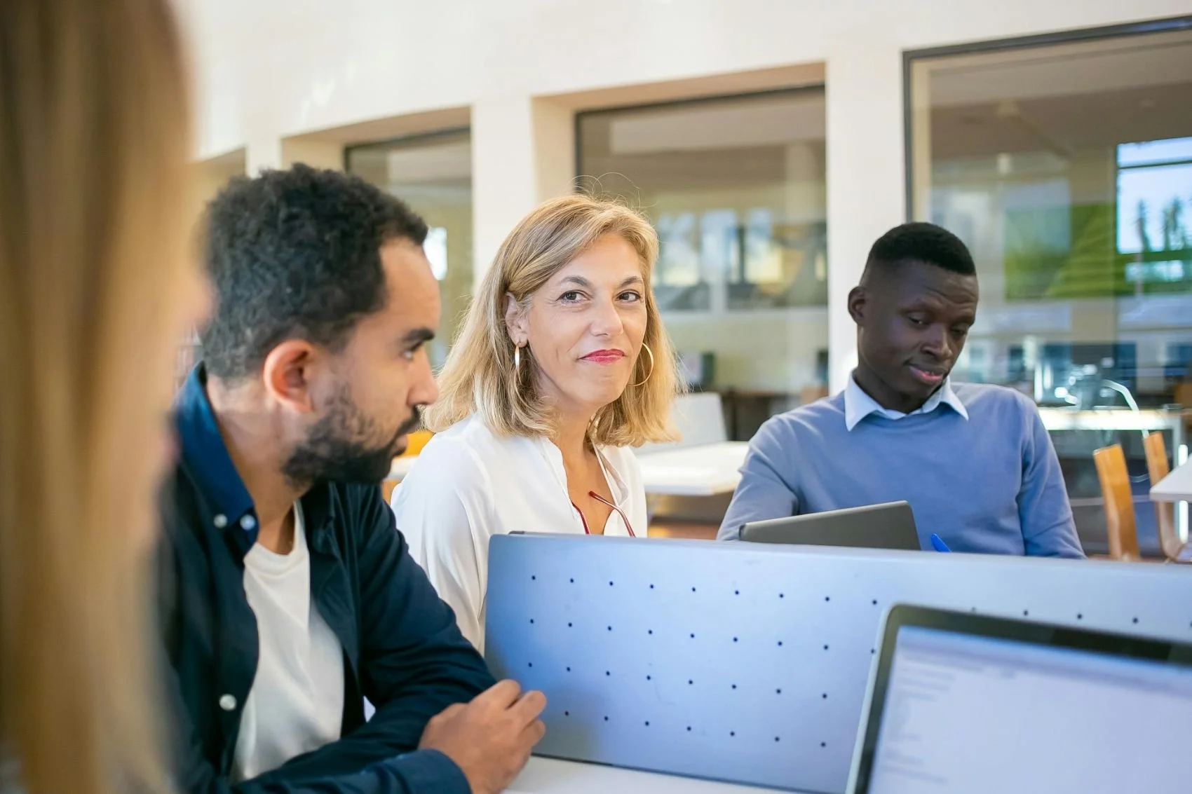 Group of four diverse professionals working together in an office, using laptops and sitting at a shared desk.