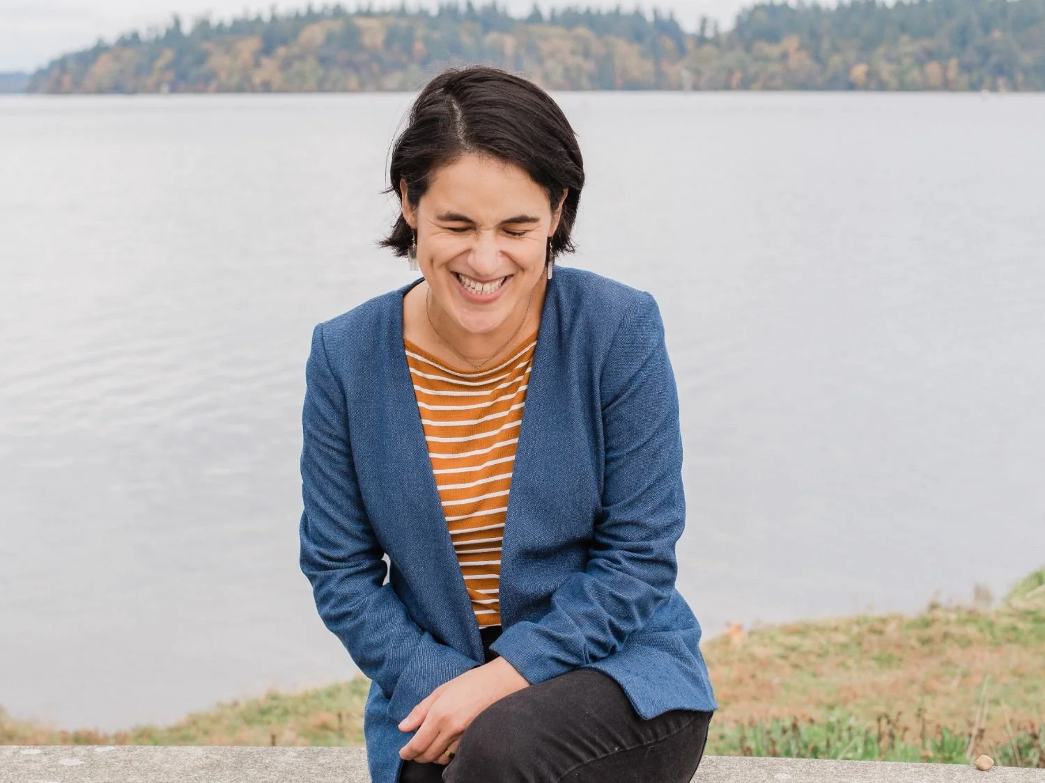 A woman with short dark hair smiling and laughing outdoors near a body of water, with trees in the background.