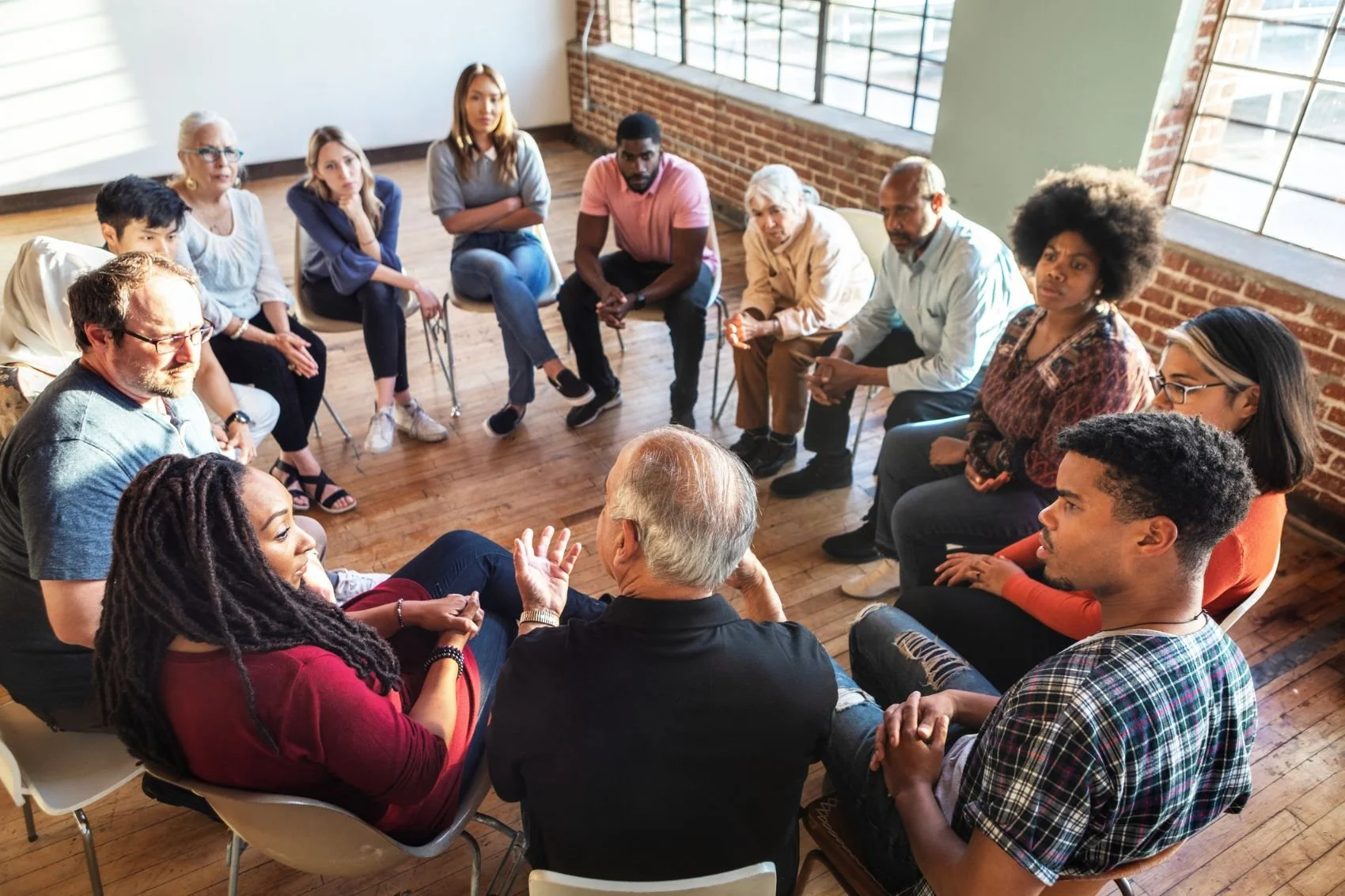 A diverse group of people sitting in a circle in a spacious room with wooden floors, brick walls, and large windows. They are engaged in a discussion or support group.