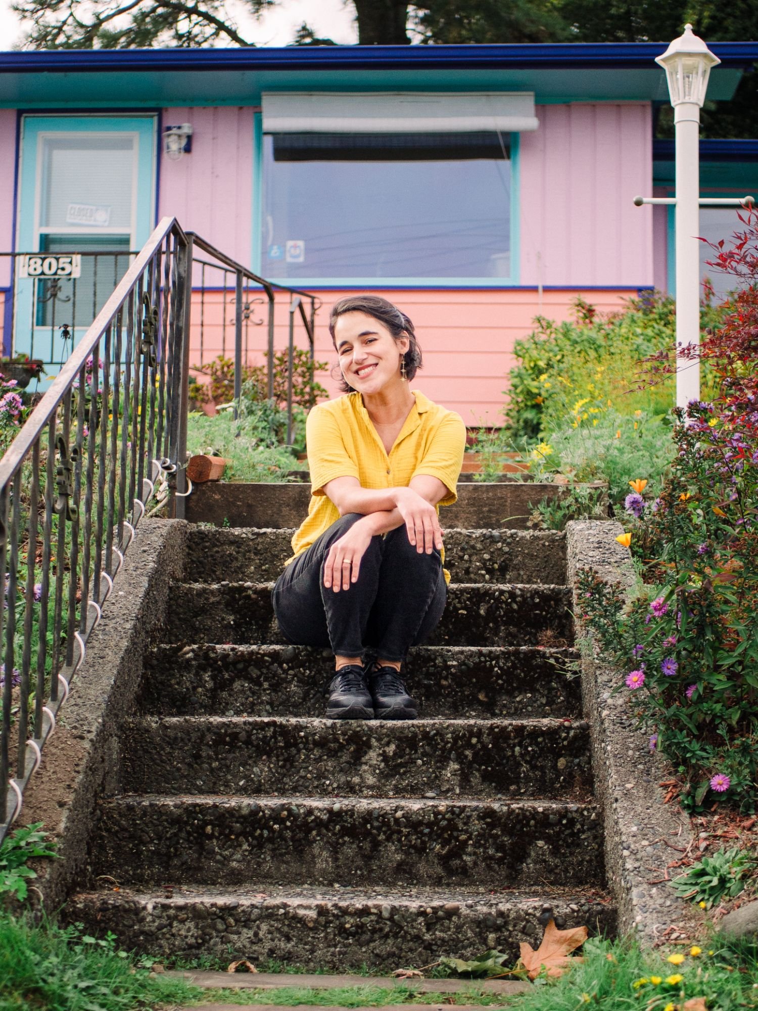 A young woman with short dark hair, wearing a yellow shirt, black pants, and black shoes, is sitting on the concrete steps of a garden in front of a pink and turquoise house. She is smiling and looking at the camera, with her arms resting on her knees.