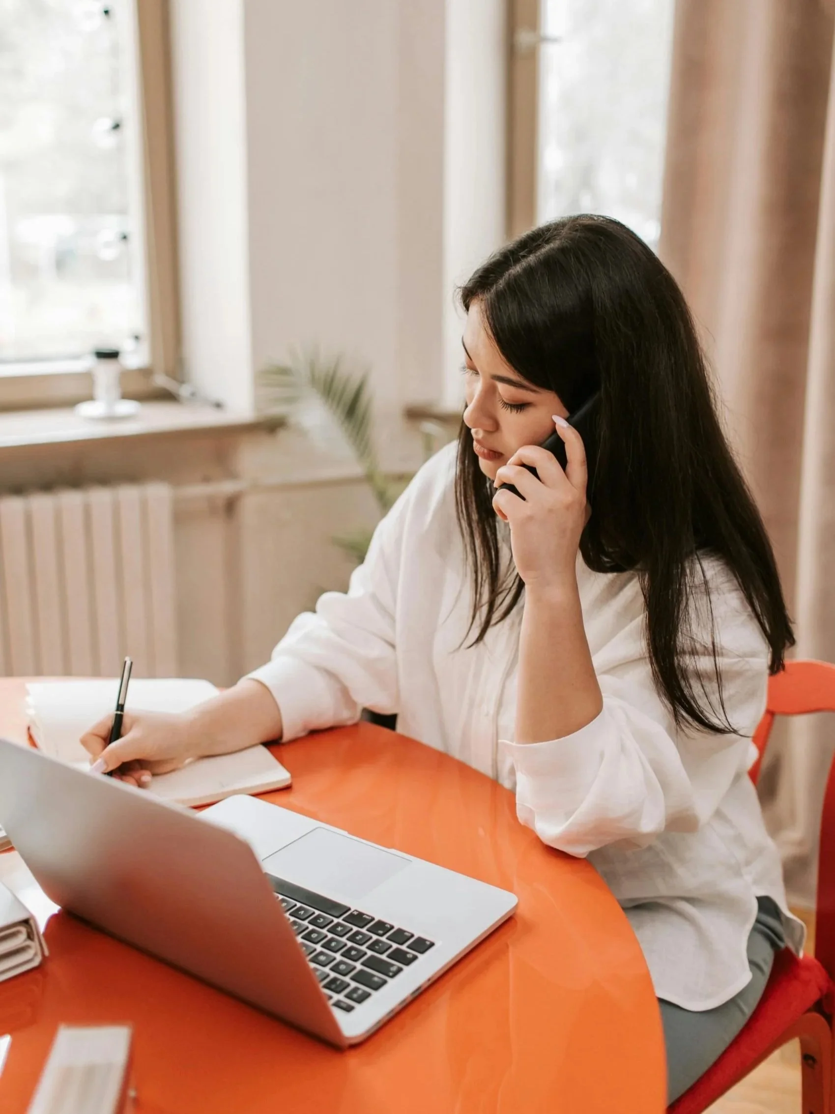 Woman with long dark hair sitting at a round orange table, talking on a landline phone, with an open laptop and a notebook in front of her, in a well-lit room with windows and curtains.