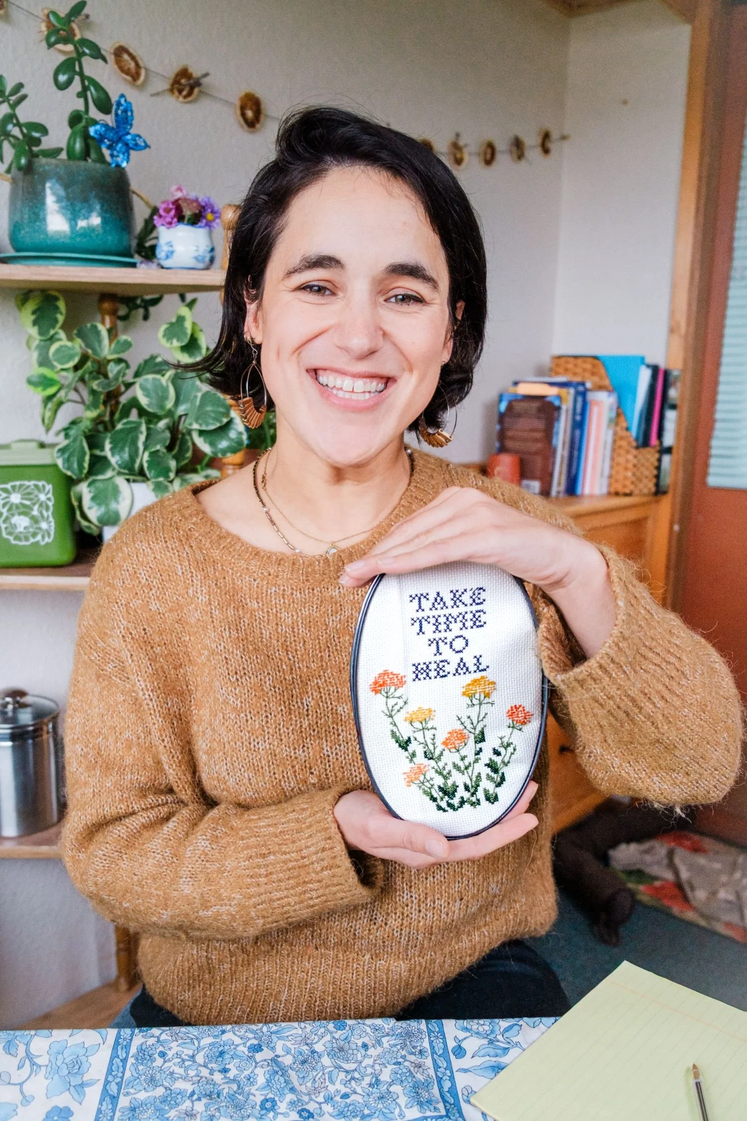 A woman with short dark hair smiling while holding an oval embroidery hoop with the words 'Take Time to Heal' and embroidered flowers.