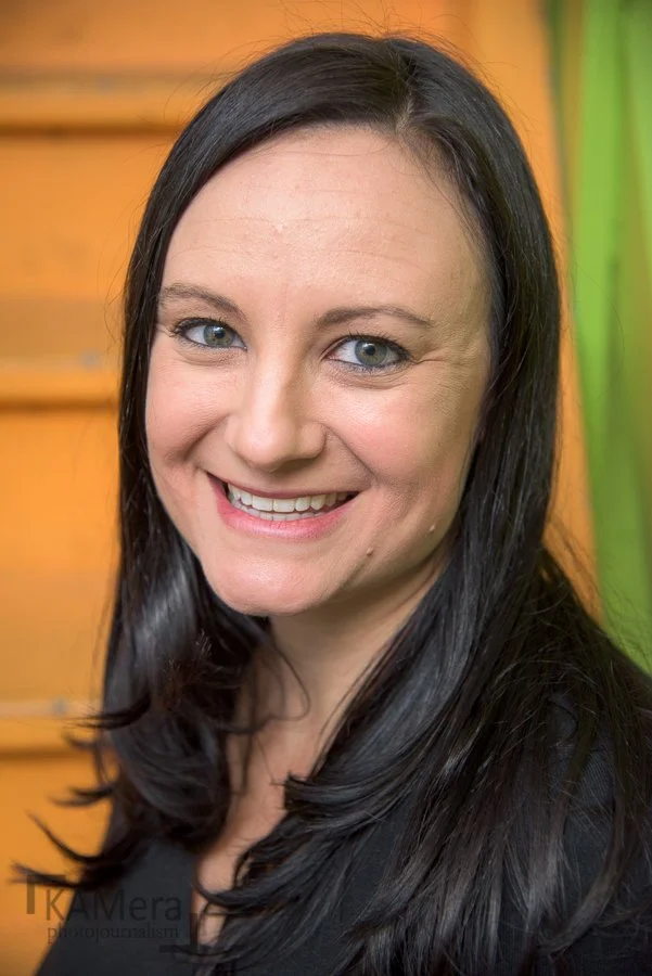 A woman with long black hair smiling, with a blurred orange and green background.