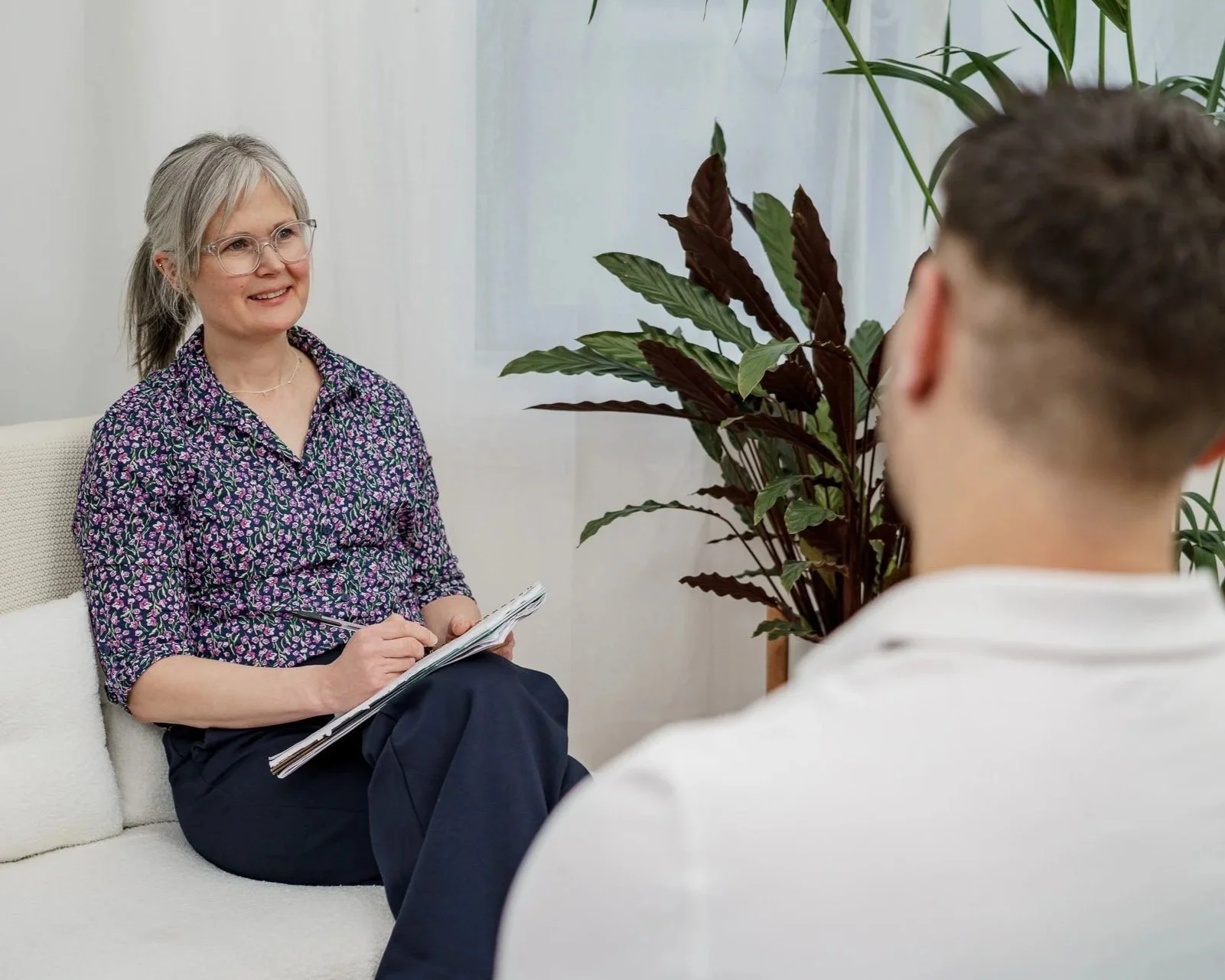 A female therapist or psychologist with gray hair and glasses smiling during a therapy session, sitting on a white couch with a notepad, in a room with beige walls and large green plants. Parent support or therapy session.