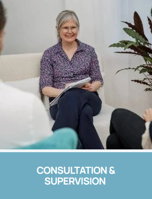 Therapist or psychologist with gray hair and glasses, wearing a floral blouse, sitting on a beige couch, holding a notebook, in a consultation or supervision with another person partially visible with a label reading "Consultation & Supervision".