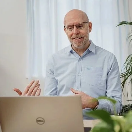 Male therapist or psychologist with glasses and a beard, dressed in a light blue button-up shirt, sitting in front of a laptop, smiling and gesturing with his hand, with a plant in the background. online therapy, supervision, consultation